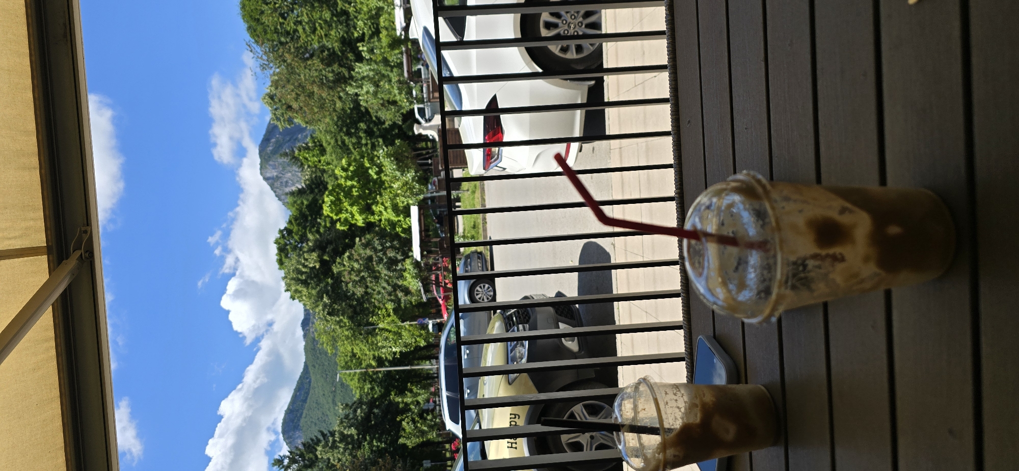 The image shows a view from a cafe or outdoor seating area, looking out towards a street and mountains. In the foreground, there are two plastic cups with straws, containing a light brown beverage. The cups are on a wooden table. A black metal railing separates the seating area from the street. Several cars are parked on the street, including a yellow car with "Ha py" written on its side. Beyond the street, there is a line of trees and then mountains under a blue sky with white clouds. The sun is shining, casting shadows.