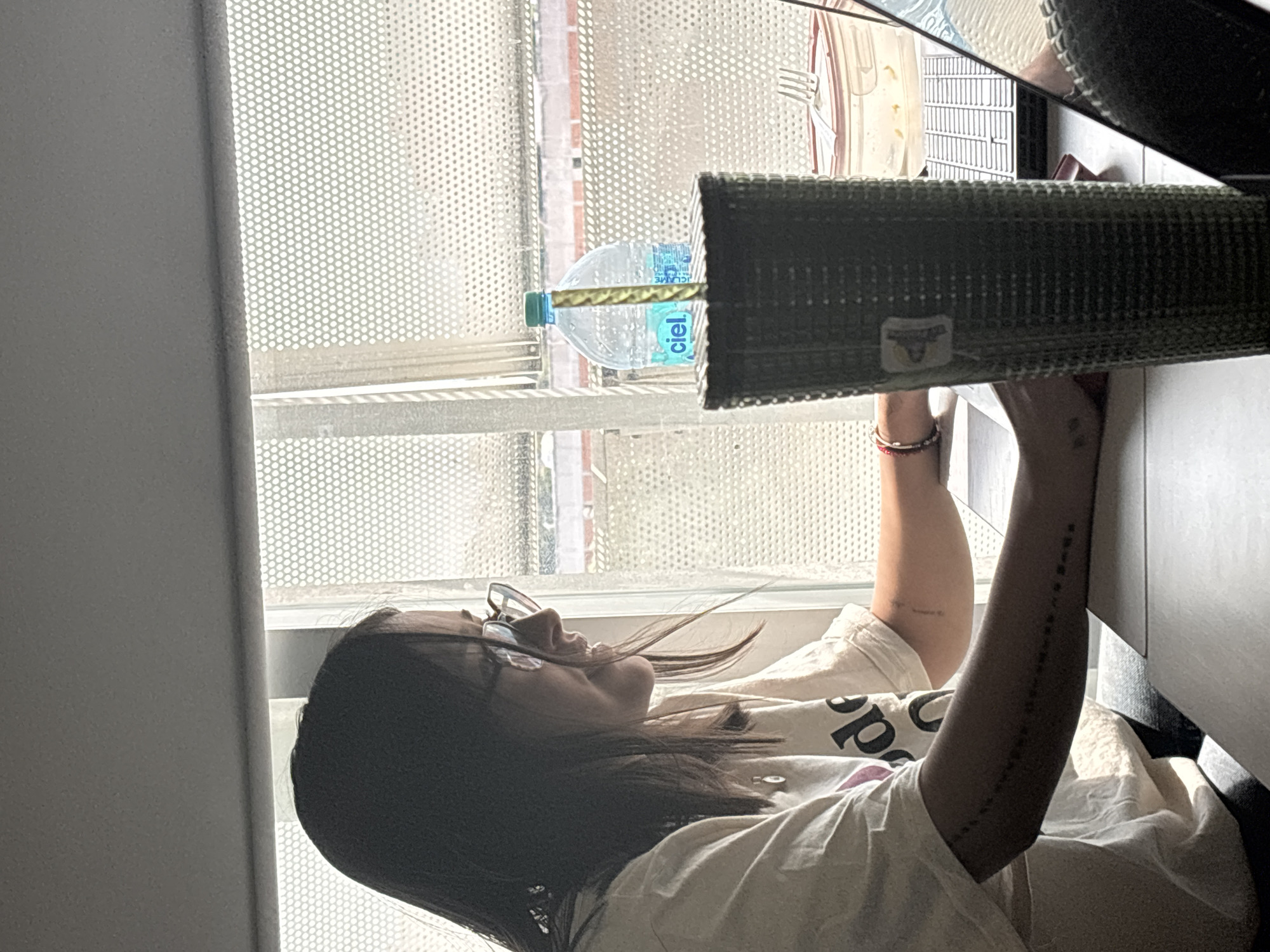 A young woman with long, dark hair and glasses is seen at an indoor location in Mexico City, Mexico. She wears a white t-shirt. She's reaching into a cabinet drawer, with a water bottle visible on the window ledge behind her. The window is covered with a mesh-like screen. The drawer appears to contain items wrapped in a green woven material. The woman's arms are raised, revealing tattoos.