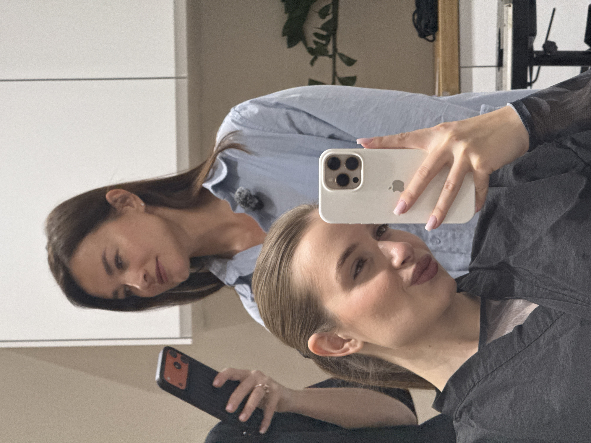 The image displays a mirror reflection of two women indoors in Budva, Montenegro. In the foreground, a woman with light brown hair, pulled back, takes a mirror selfie using a white iPhone. She holds the phone with her right hand, showcasing her light pink manicured nails and the phone's triple camera system and Apple logo. Her facial expression is a playful "duck face" or kissy face, and she wears a dark grey or black top, possibly with a sheer shoulder detail.
Behind her and to the left, also seen in the reflection, is another woman with long dark hair, wearing a light blue button-up shirt. A small dark microphone or clip is attached to her collar. She looks downwards with a neutral expression, holding a dark smartphone with an orange accent on its back in her left hand.
The reflected background includes light-colored walls or cabinets and a dark green plant on the upper left side, suggesting an indoor environment. The scene captures a casual moment, possibly during preparations or a break, with the reflection providing a unique perspective. No specific time of day or weather is discernible from the indoor setting.