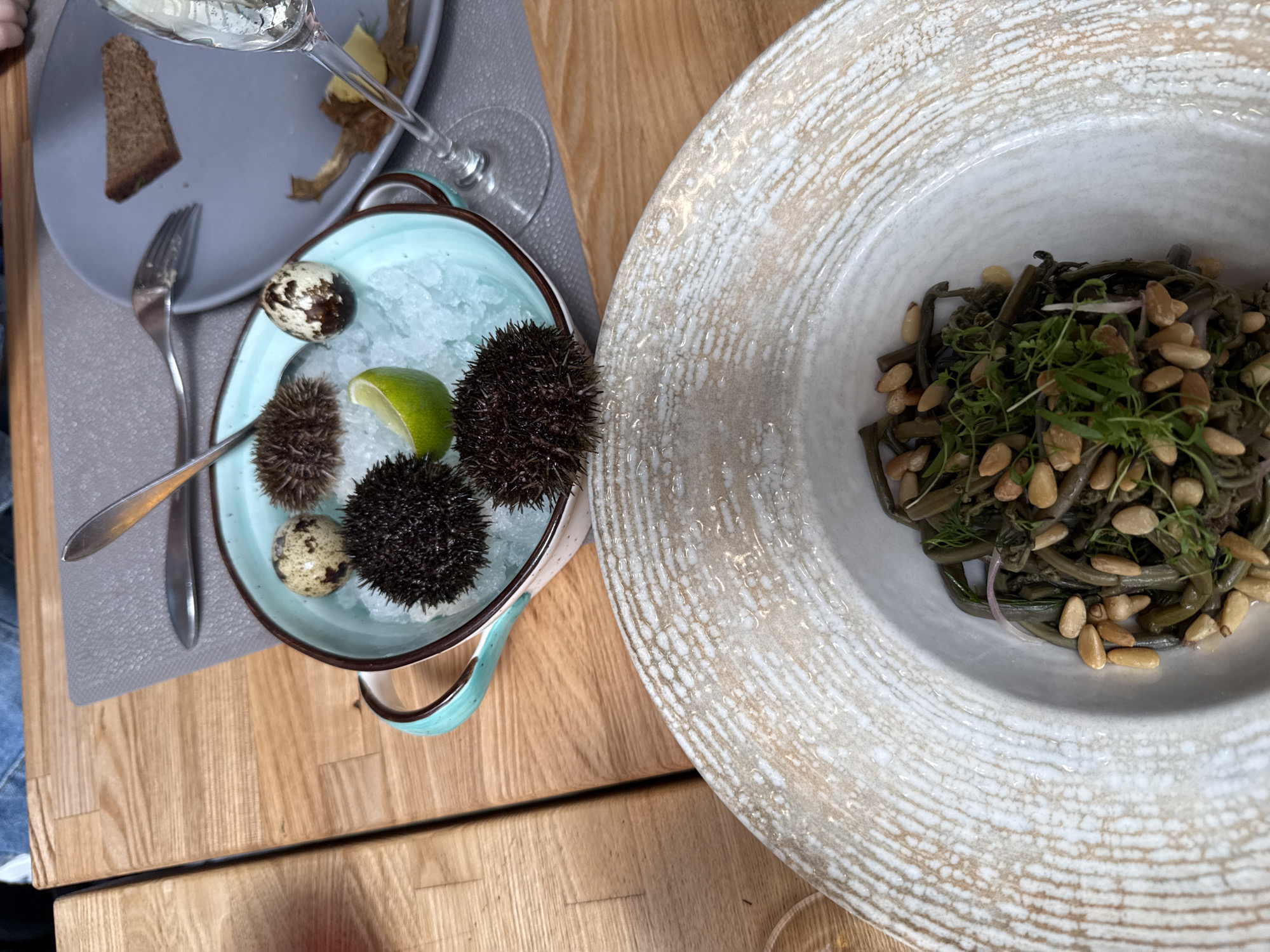 The image shows a table setting with two dishes, likely in Russky, Russia. On the left is a small, light blue bowl filled with ice, sea urchins, quail eggs, and a lime wedge, accompanied by a fork. Next to it is a plate with bread and some fried food. On the right, a large, shallow white bowl contains a dish of dark green seaweed salad with pine nuts and herbs. The table has a light brown, wood-grain surface. The overall setting suggests a meal at a restaurant, possibly specializing in seafood.