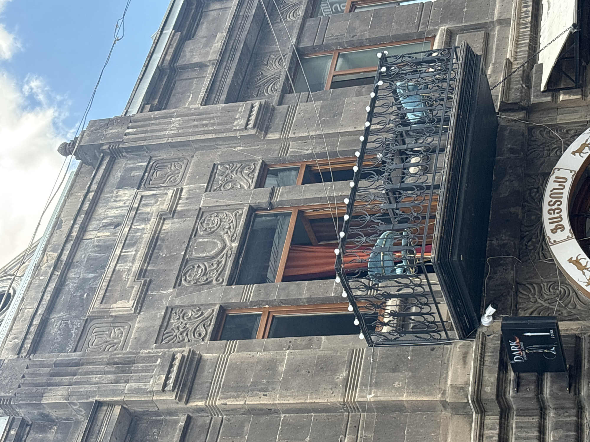This is a close-up, angled shot of an old stone building with intricate carvings. The building has multiple windows, some with wooden frames, and an ornate metal balcony. The balcony is decorated with what appear to be small, round lights. Inside the balcony, a person wearing a blue garment is visible. The sky above is partly cloudy and appears blue. A security camera is mounted on the building near a black sign with white lettering that reads "DARK HOOKAH LOUNGE" and an arrow pointing to the right. To the right of the sign, a circular sign with an ornate design and text in a non-Latin alphabet is partially visible. The overall impression is that of an old, possibly European, urban setting. The lighting suggests it is daytime.
