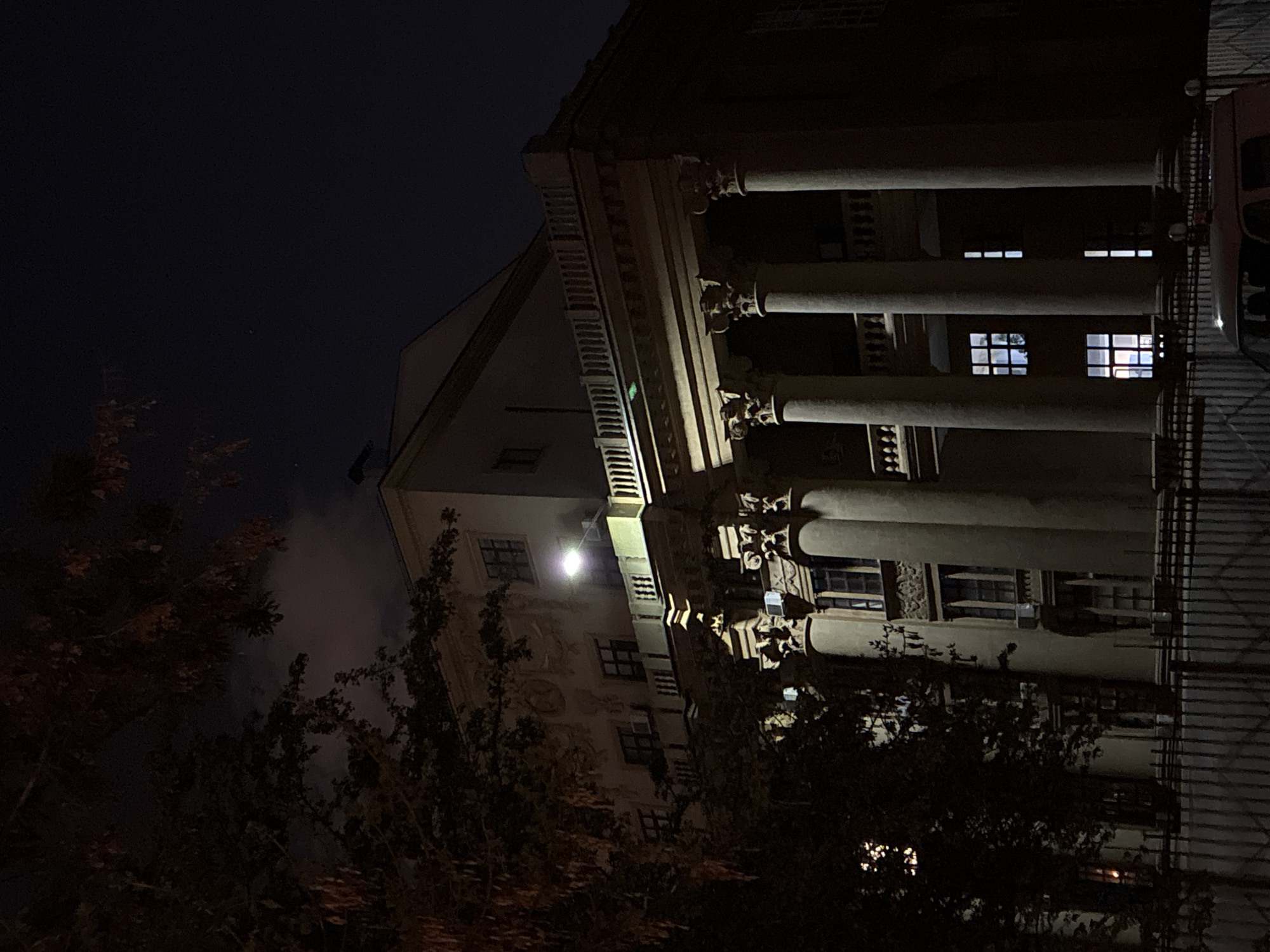 The image shows a multi-story building at night in Moscow, Russia. The architecture features classical elements such as columns and ornate detailing. Illuminated windows are visible. Trees partially obscure the lower floors. The sky is dark. A vehicle is partially visible on the right side of the frame.