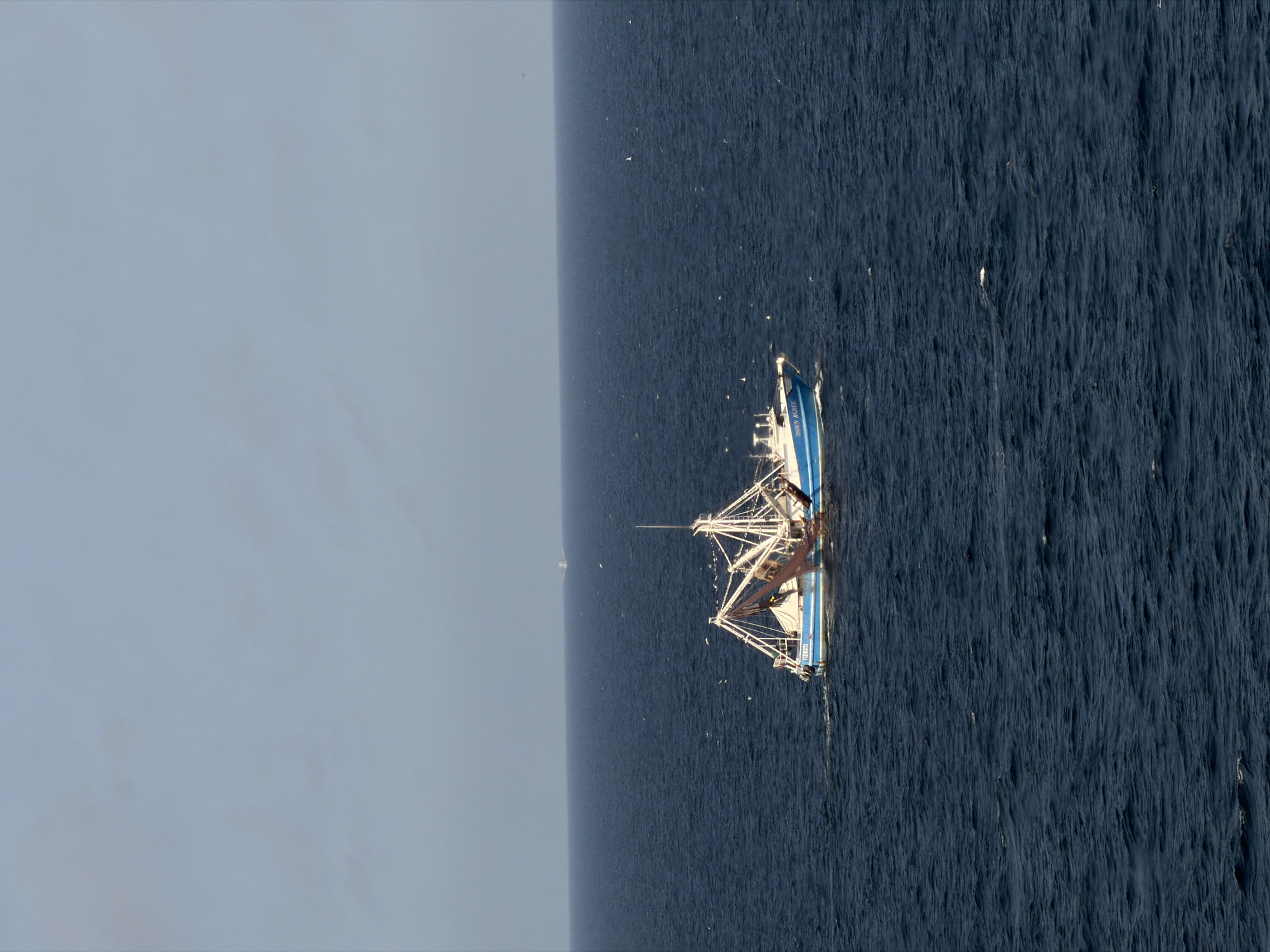 The image, taken from an elevated perspective, shows a fishing vessel in the open sea under a hazy sky. The vessel, appearing to be a trawler or shrimper, has a prominent blue hull with a white superstructure and deck. It is equipped with extensive fishing gear, including large booms extended outwards, rigging, and visible nets, suggesting it is either actively fishing or prepared to do so.
On the bow of the vessel, the name "SHIRLEY MARIE" is clearly visible. On the side of the hull, the text "USCG 1128311" (or similar numerals) can be read.
The water is a deep, dark blue, with a textured and choppy surface indicating movement or waves. Patches of white foam and small whitecaps are visible around the vessel and scattered across the water. The sky is a uniform, light blue-grey, suggesting an overcast or hazy daytime condition. In the distant horizon, a faint, indistinct structure, possibly another vessel or an offshore platform, can be seen. Several small white specks, possibly birds or debris, dot the water surface. No specific individuals or their emotions are discernible.