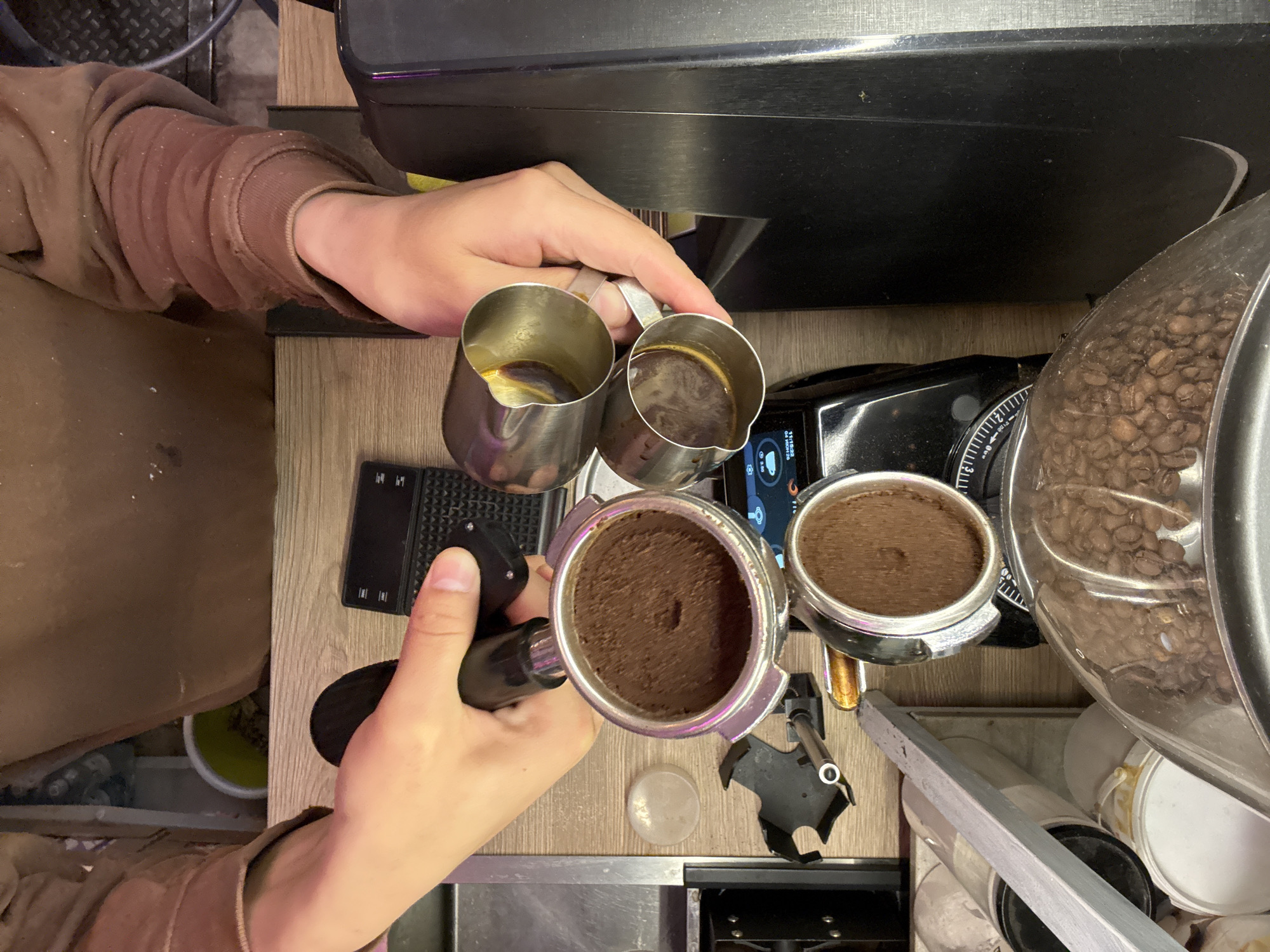 A high-angle, vertical shot captures a person's hands actively engaged in preparing espresso on a light wood-grain countertop, likely in a home kitchen or a small coffee bar in Moscow, Russia.
The primary subjects are the person's hands and several coffee-making tools. The right hand holds two small, stainless steel pitchers, each containing freshly pulled espresso shots with visible dark liquid and some crema. The left hand firmly grips the handle of a double-spout espresso portafilter, which is packed level with finely ground coffee. Another similar portafilter, also filled with ground coffee, rests on a black surface to the right, suggesting it's either ready for the machine or has just been ground.
Parts of black coffee equipment are visible: an espresso machine's top section to the upper right and a coffee grinder below it, featuring a clear hopper brimming with whole coffee beans. A small digital display on the grinder shows "11:17:32" and "04/00/23," along with a coffee cup icon. To the left, a black, textured rubber mat is placed on the counter. Scattered specks, possibly coffee grounds, are visible on the brown sleeve of the person's garment and the countertop.
The activity depicts a precise stage in the espresso-making process, showcasing both extracted shots and fresh grounds, indicating a focused and detailed approach to coffee preparation.