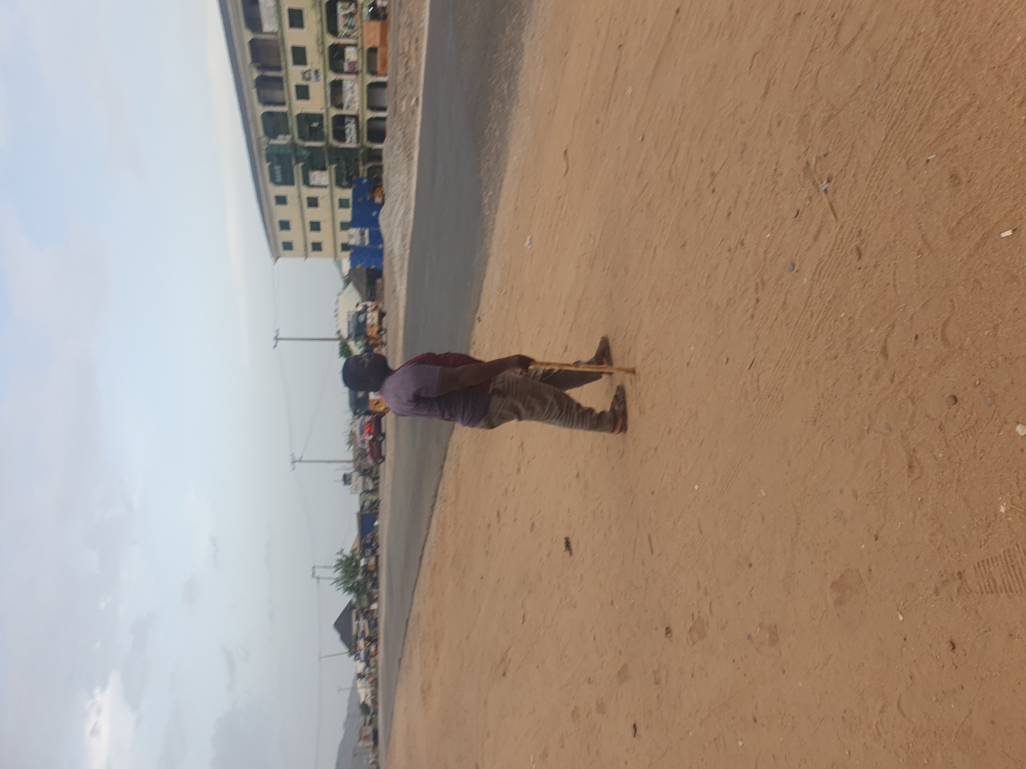The image captures an outdoor street scene in Lokoja, Nigeria, under an overcast or hazy daytime sky. The composition is dominated by a reddish-brown dirt or sand path in the foreground and midground, running parallel to a paved road on its left.
A man is standing on the dirt path, with his back mostly to the viewer, angled slightly to the right. He is wearing a light purple short-sleeved shirt and light-colored trousers. He holds a long, thin, light-colored stick or staff vertically, with its end resting on the ground near his right foot.
To the left of the paved road, in the background, a multi-story building with a light-colored facade and numerous dark windows is visible. Further down the road, smaller buildings, blurry vehicles, and several utility poles with overhead wires line the street. The dirt path shows subtle indentations and scattered small debris. No discernible text is visible anywhere in the frame.