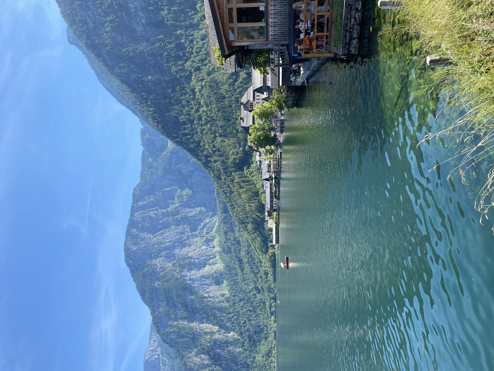 This image captures a serene waterside scene in Hallstatt, Austria, on a clear, sunny day. The foreground features the calm, teal-colored waters of Hallstätter See, with visible ripples and sunlight reflecting on its surface. To the right, a portion of a wooden building with a covered patio extends over the water, where several individuals are seated at tables, some appearing to be dining or conversing. Beyond this structure, the picturesque town of Hallstatt unfolds along the lake's edge, characterized by traditional Austrian architecture with pitched roofs and pastel-colored facades, nestled against a backdrop of lush, green mountains. A red boat with a single occupant and a parasol is visible on the lake, further enhancing the tranquil atmosphere. The background is dominated by steep, imposing mountains covered in dense evergreen forests, with rocky outcrops visible on their slopes, all under a bright blue sky with sparse wisps of clouds. The overall impression is one of natural beauty and idyllic village life.
