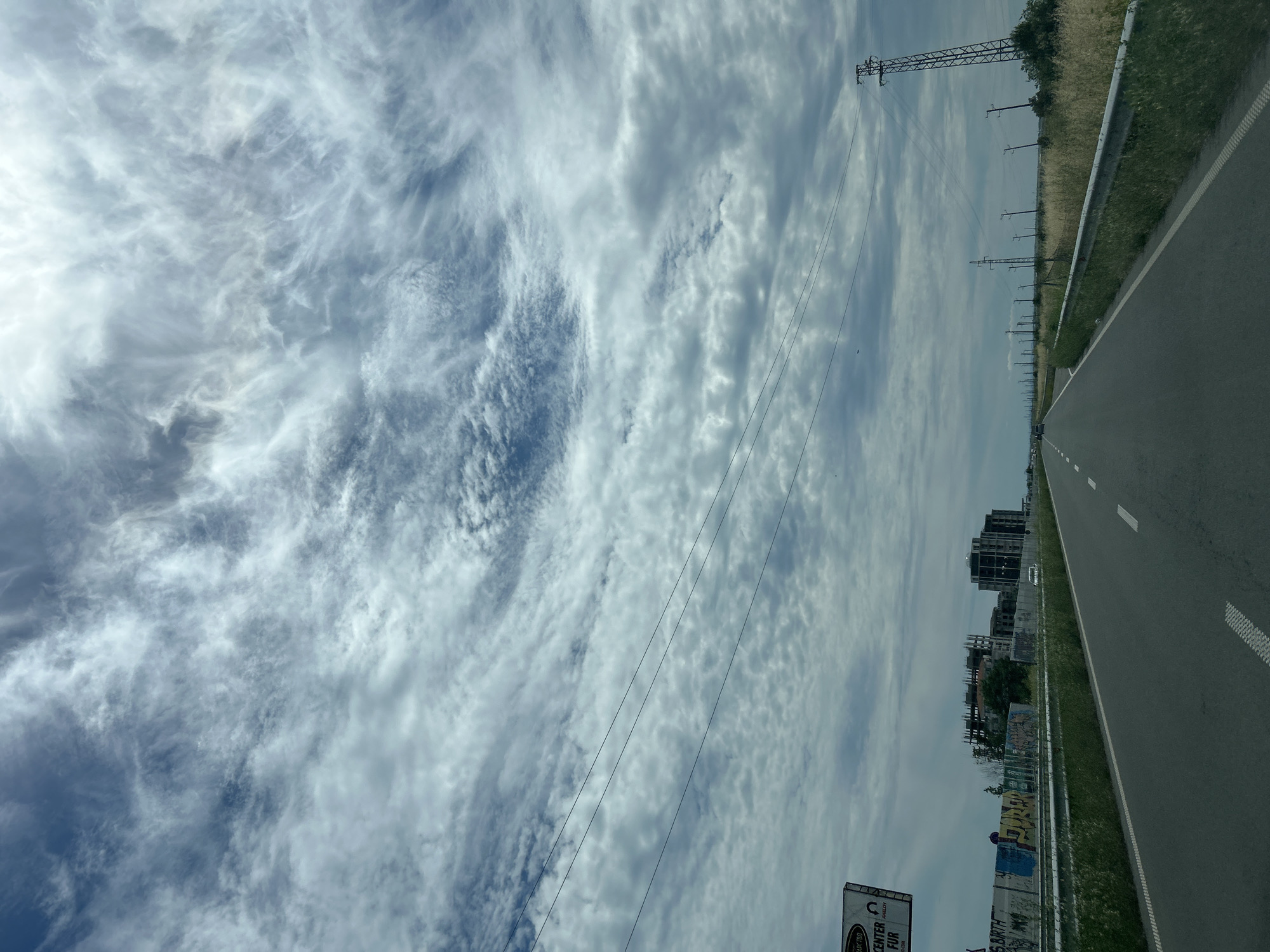 The media file depicts a roadside view under a cloudy sky, oriented vertically. The scene looks out over a paved road with white lane markings, suggesting a highway or major road. To the right of the road, there is a grassy verge and a metal guardrail. Beyond the guardrail, a field of tall, dry grass stretches towards a line of power poles and electrical wires that extend into the distance. In the background, partially obscured by the power lines, are several buildings, some appearing to be under construction with visible scaffolding. Graffiti is visible on a low wall or barrier closer to the foreground. The sky is filled with a dynamic pattern of white and gray clouds, with some patches of blue visible. The sun is not directly visible, but the brightness of the clouds suggests it is daytime, likely mid-morning or afternoon. The overall atmosphere is one of travel and passing through a semi-urban or developing area. There is a sign visible in the lower right corner with some text, but it is partially cut off and illegible. Another sign is visible near the bottom center, also partially obscured, with some legible text.