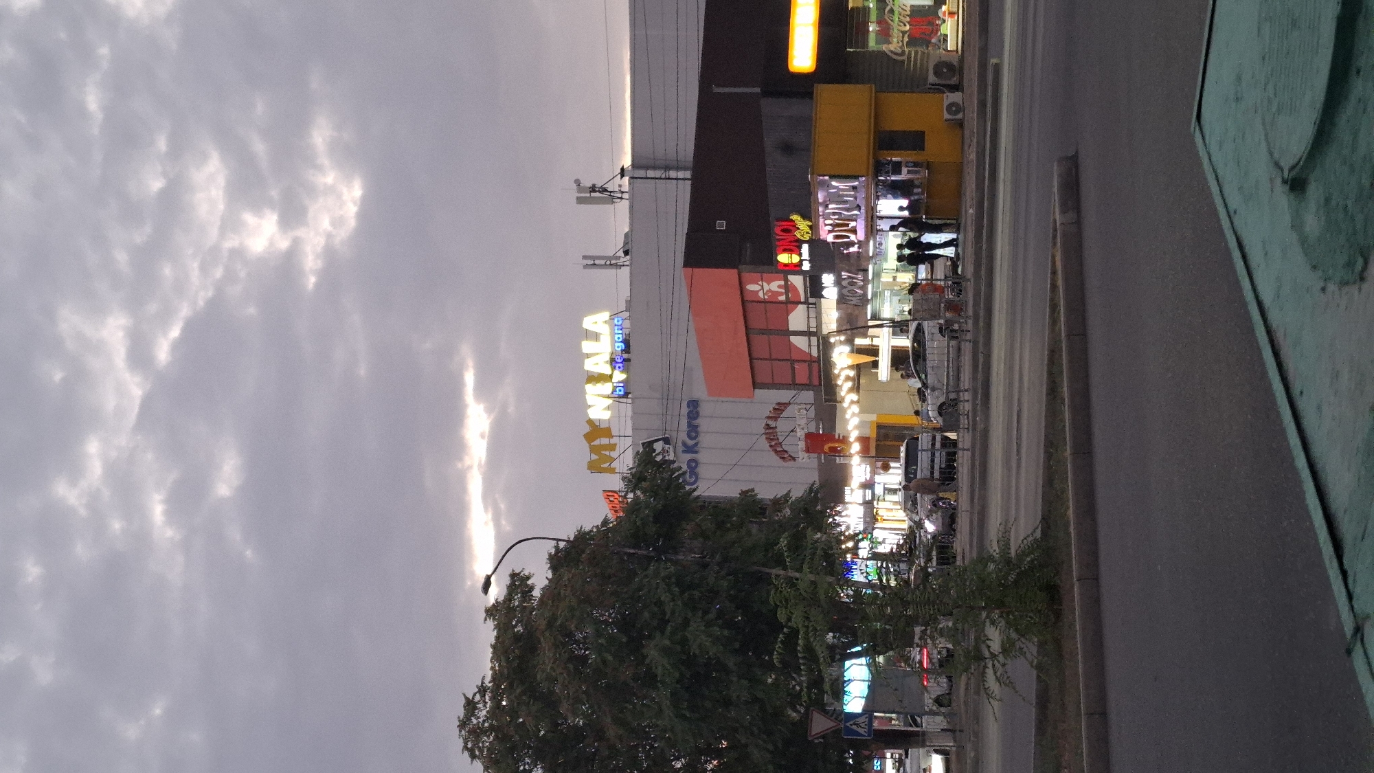 The image shows a street scene in Shymkent, Kazakhstan, at dusk. In the background, a large building with a prominent "MY NI ALA" sign is visible. Below this, a sign reads "Go Korea". Several smaller shops and businesses line the street, with illuminated signs including "Duyum" and a Coca-Cola advertisement. Cars are parked and driving on the road, and a few people are standing outside the establishments. A tree and a street lamp are on the left side of the frame. The sky is overcast with visible clouds.