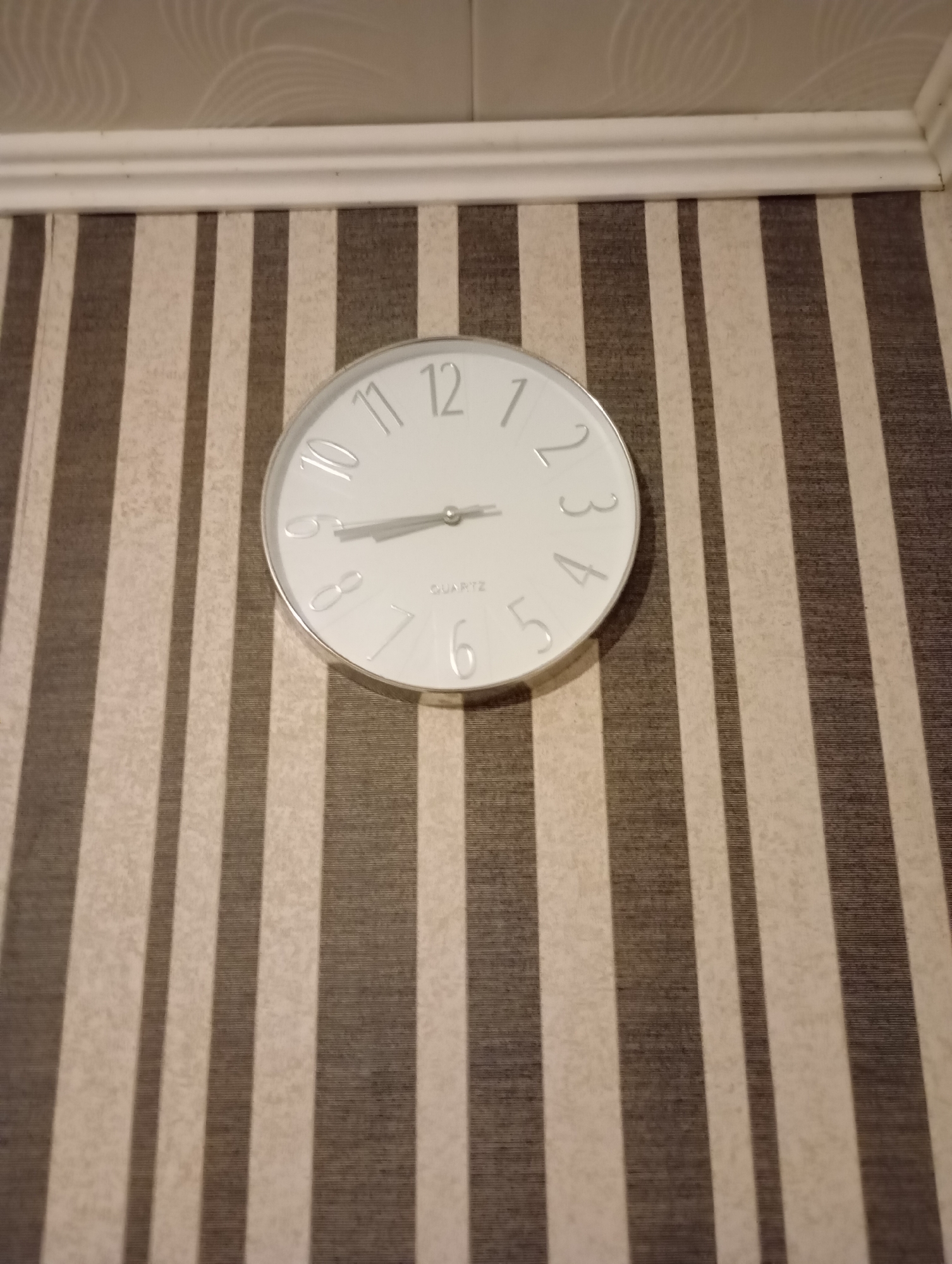 A round, white clock with silver numbers and hands hangs on a wall covered in vertical stripes of beige and brown. The clock's hour hand points to the number 9, and the minute hand points to the number 11. The wallpaper has a textured, matte finish. Above the wallpaper is a white molding. The scene is indoors, with no indication of the specific city or location beyond it being "Unknown".