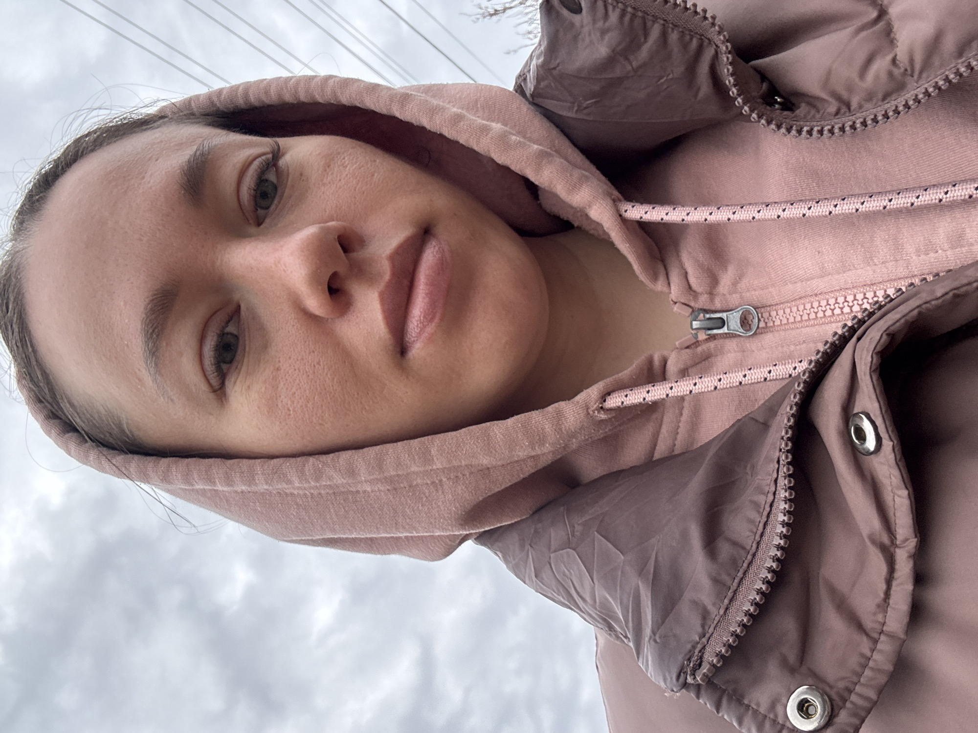 A woman is wearing a mauve hoodie and jacket, looking slightly down at the camera. The background shows an overcast sky and power lines. The location is inferred to be Ферма, Russia.