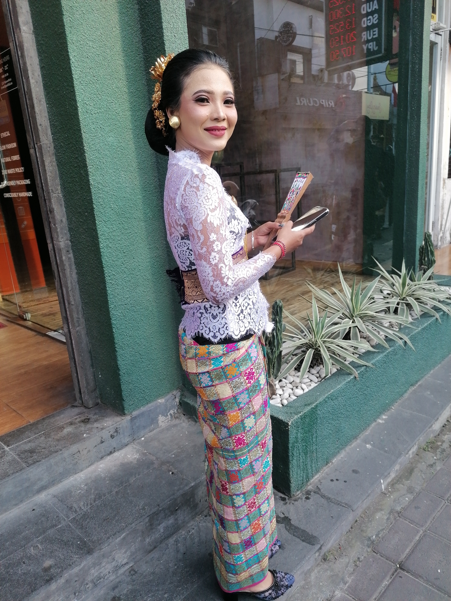 A young Indonesian woman, adorned in traditional Balinese attire, stands outdoors near a building with a green textured facade. She wears a white lace top with intricate patterns and a colorful, intricately woven sarong that reaches to her ankles. Her hair is styled in an updo with a golden ornament. She is holding a folding fan and a smartphone in her hands. The background features a shop window with reflections of currency exchange rates and shop signs. A planter box with several small plants and white pebbles is situated in front of the building. The ground is paved with concrete and stone slabs. The lighting suggests it is daytime. The location appears to be a street in Ubud District, Indonesia, indicated by the cultural attire and likely a commercial area.