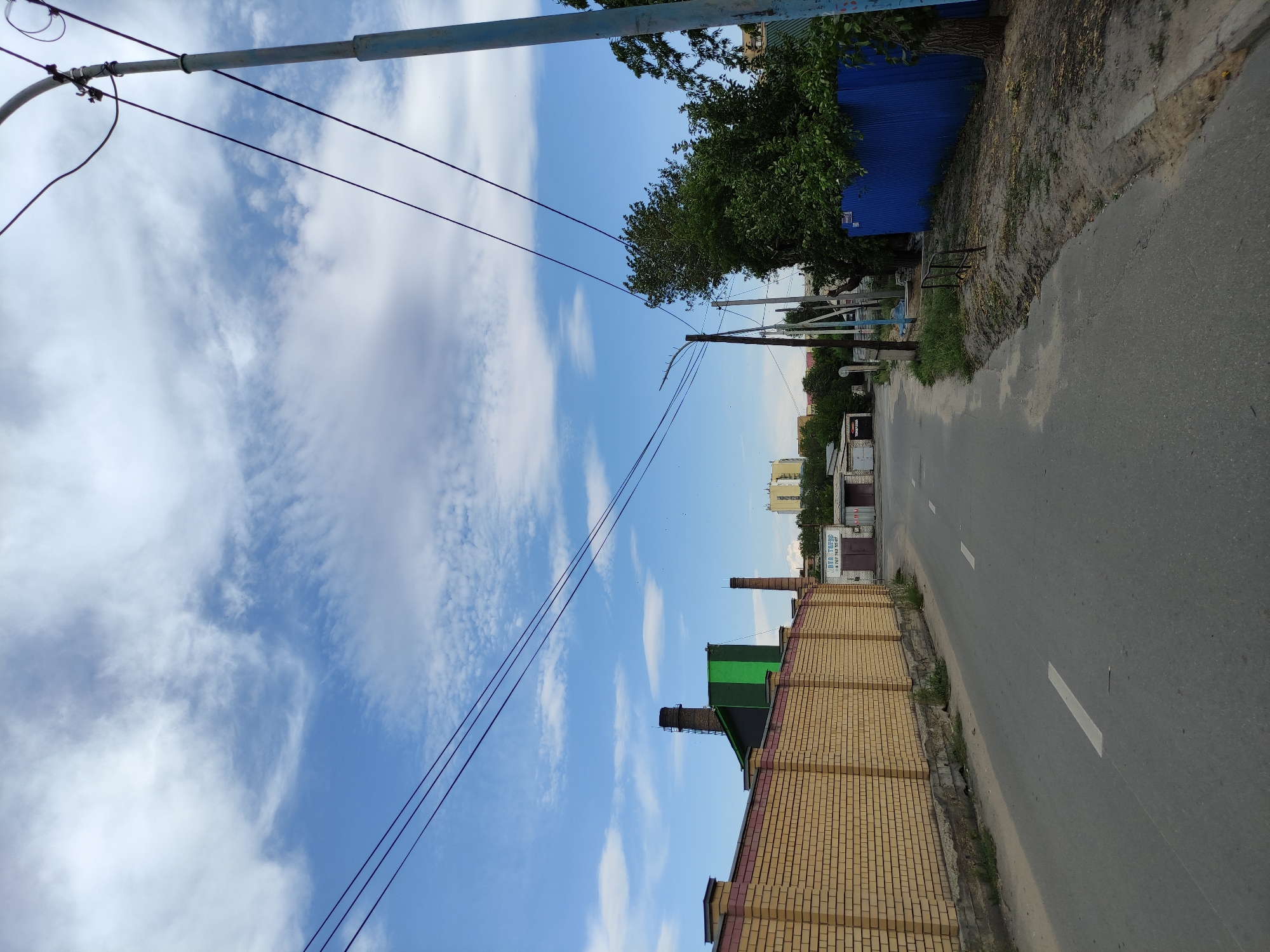 The image captures a street scene in Semey, Kazakhstan, under a partly cloudy sky. On the left, a yellow brick wall with a red-brown cap lines the street, separating it from an industrial-looking area with tall chimneys, one of which has a green structure attached. In the distance, a multi-story building is visible. The road itself is asphalt with white lane markings and a white line along the side. On the right side of the road, a tall blue metal fence is prominent, with lush green trees behind it. A power pole with overhead wires stands on the right edge of the frame, and another pole with a street light is visible further down the street on the right. The ground on the right side of the road appears to be a mix of dirt and sand. The overall impression is an urban or semi-industrial area.