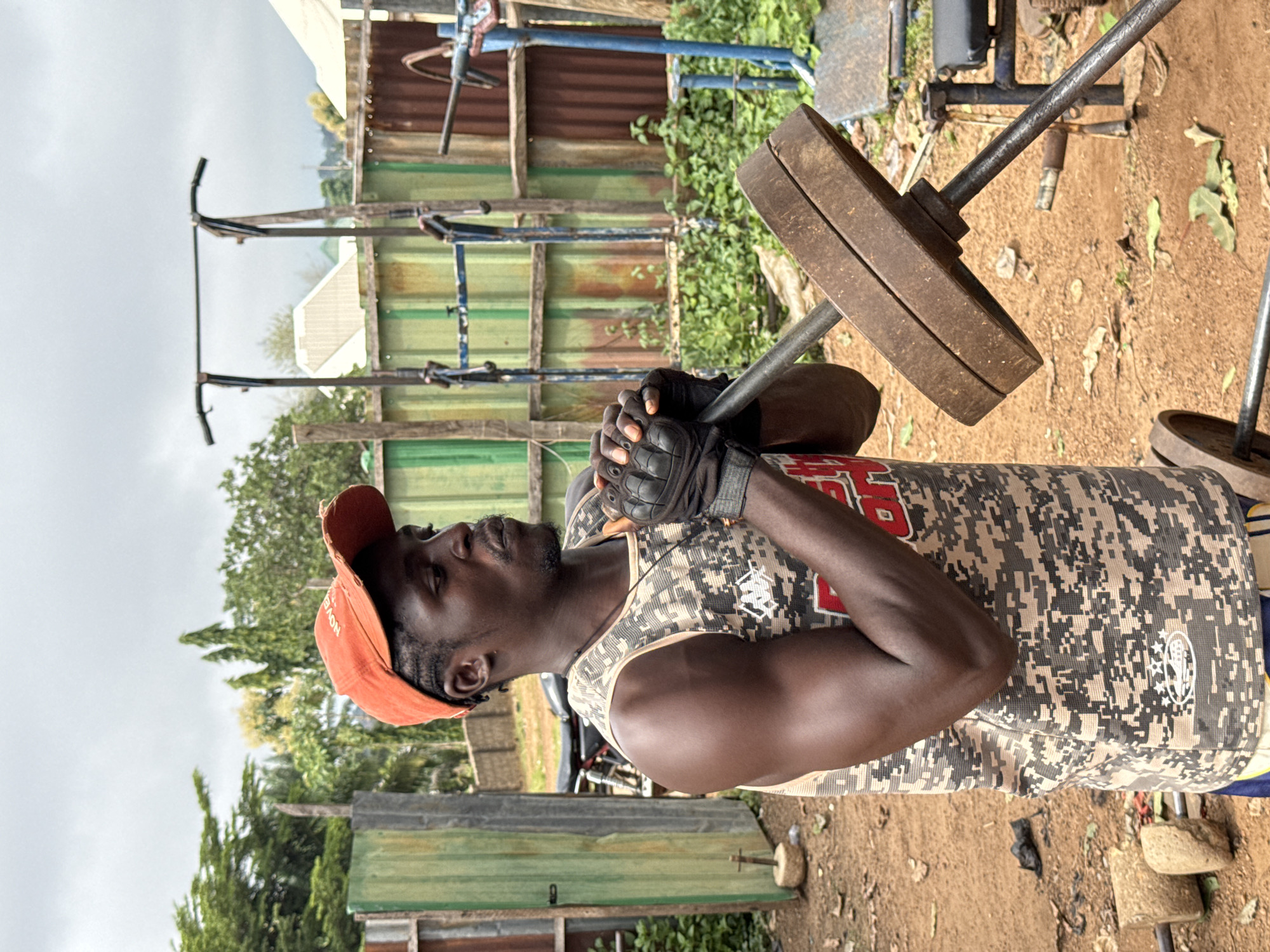 The image shows a man lifting weights outdoors in Akwanga, Nigeria. He is wearing an orange baseball cap, fingerless workout gloves, and a camouflage tank top. The man is focused and appears to be exerting himself as he lifts a barbell with weights. The background includes a makeshift outdoor gym with various pieces of equipment, including a bench and other weights. There are also corrugated metal structures, vegetation, and trees, suggesting a somewhat rural or informal setting. The ground is dirt and scattered with leaves. The lighting indicates it is daytime, and the sky is overcast. There is no discernible text in the image.