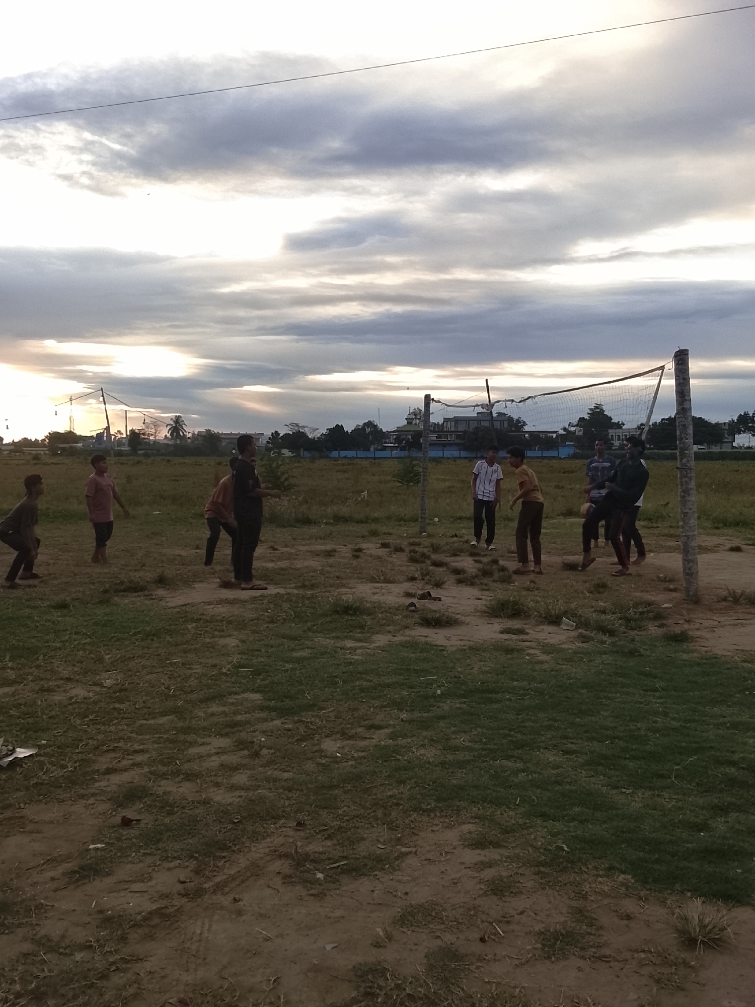 In the city of Dayah Baroh, Indonesia, under a cloudy sky with dramatic light filtering through, a group of young men is engaged in a game of volleyball on an open, grassy field. The court is marked by a net suspended between two wooden posts. The players, dressed in casual clothing, are actively participating, with some in mid-action, jumping or preparing to hit the ball. The surrounding environment is somewhat unkempt, with patches of dry grass and dirt. In the background, trees and some buildings are visible, suggesting an outdoor recreational area on the outskirts of a settlement. The overall scene depicts a moment of leisure and sport amidst a natural, undeveloped landscape.