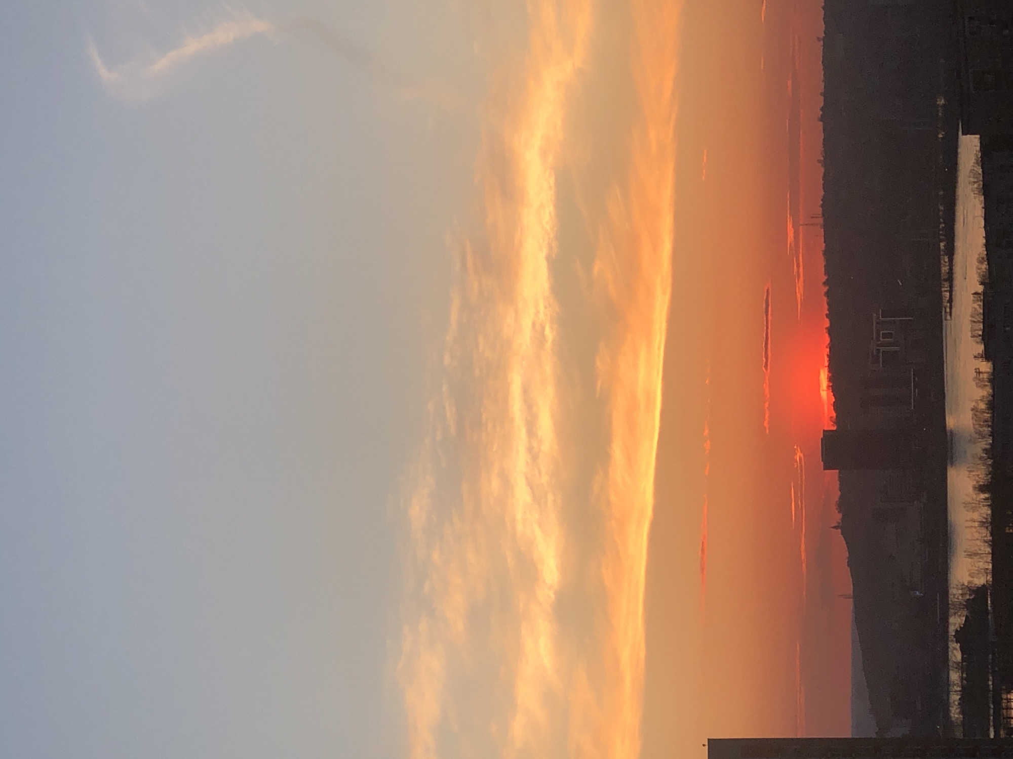 The image shows a sunset over the city of Krasnoyarsk, Russia. The sky is a gradient of colors, transitioning from light blue to orange and red. The sun appears as a bright red disk near the horizon, obscured by buildings. Silhouetted buildings and trees are visible against the colorful sky. A body of water reflects the sunset colors. The image is rotated.