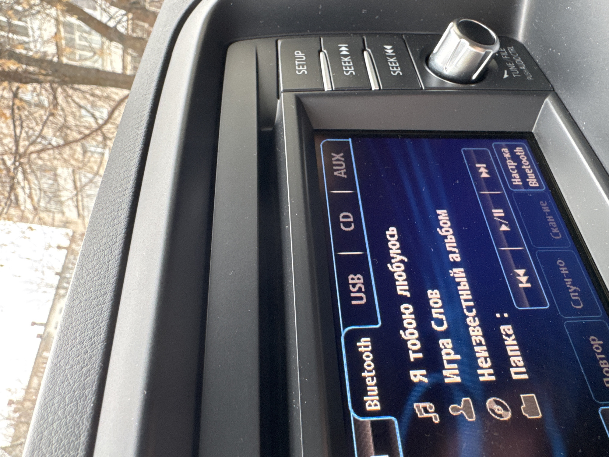 The image shows the infotainment system of a car, displaying Russian text and music-related icons. The screen indicates that music is being played via Bluetooth from a USB source. The car's interior is visible, with buttons for "SETUP", "SEEK," "CD," and "AUX." A silver knob is present for tuning and audio control. The exterior view shows bare trees and a building, suggesting an urban environment. The Russian text on the screen implies a location in Russia, specifically Chaikovsky, as the text is in Russian.