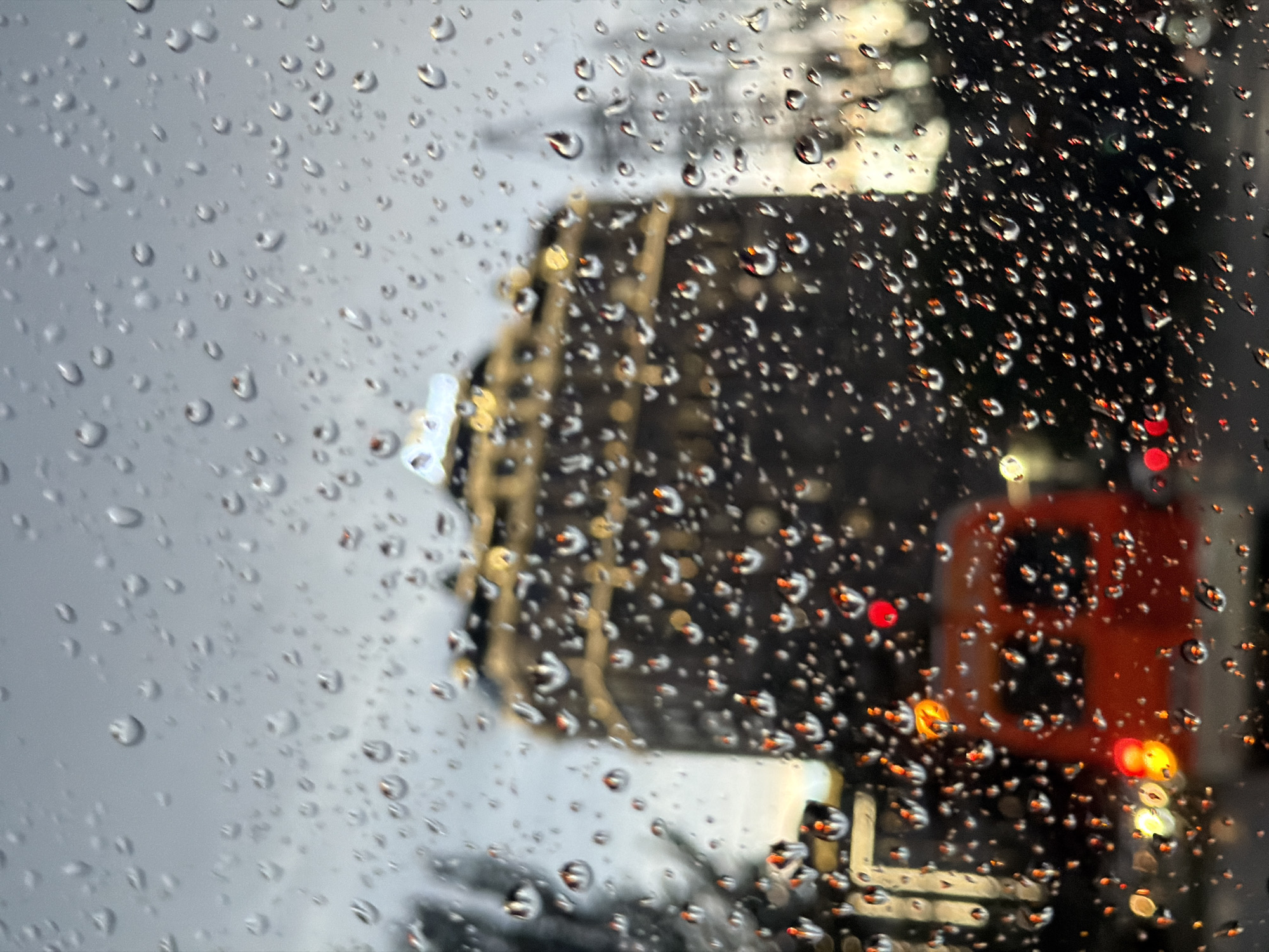 A rain-covered window obscures the view of Bishkek City, Kyrgyzstan. Through the droplets, a blurred building stands to the left, while a bus, identifiable by its rectangular shape and illuminated lights, is on the right. The background is generally gray and overcast.