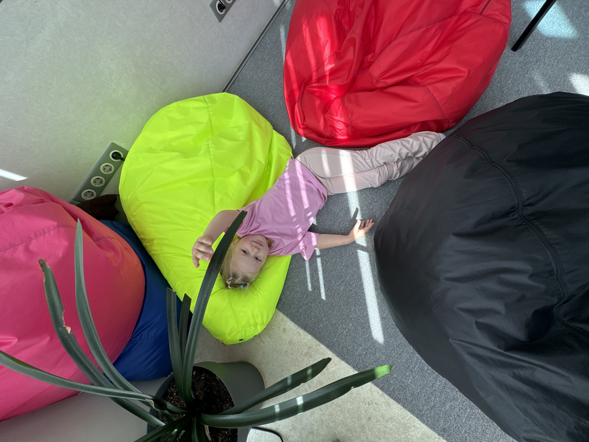 A child lies on a lime green beanbag in a room in Perm, Russia. The child is light-skinned with blonde hair and is wearing a light purple t-shirt and light tan pants. The beanbag is one of four that are arranged in the room, along with a plant in a pot. The other beanbags are red, black, and pink. The floor is gray and the walls are off-white. There is an electrical outlet on the wall near the lime green beanbag. Natural sunlight streams through the window.