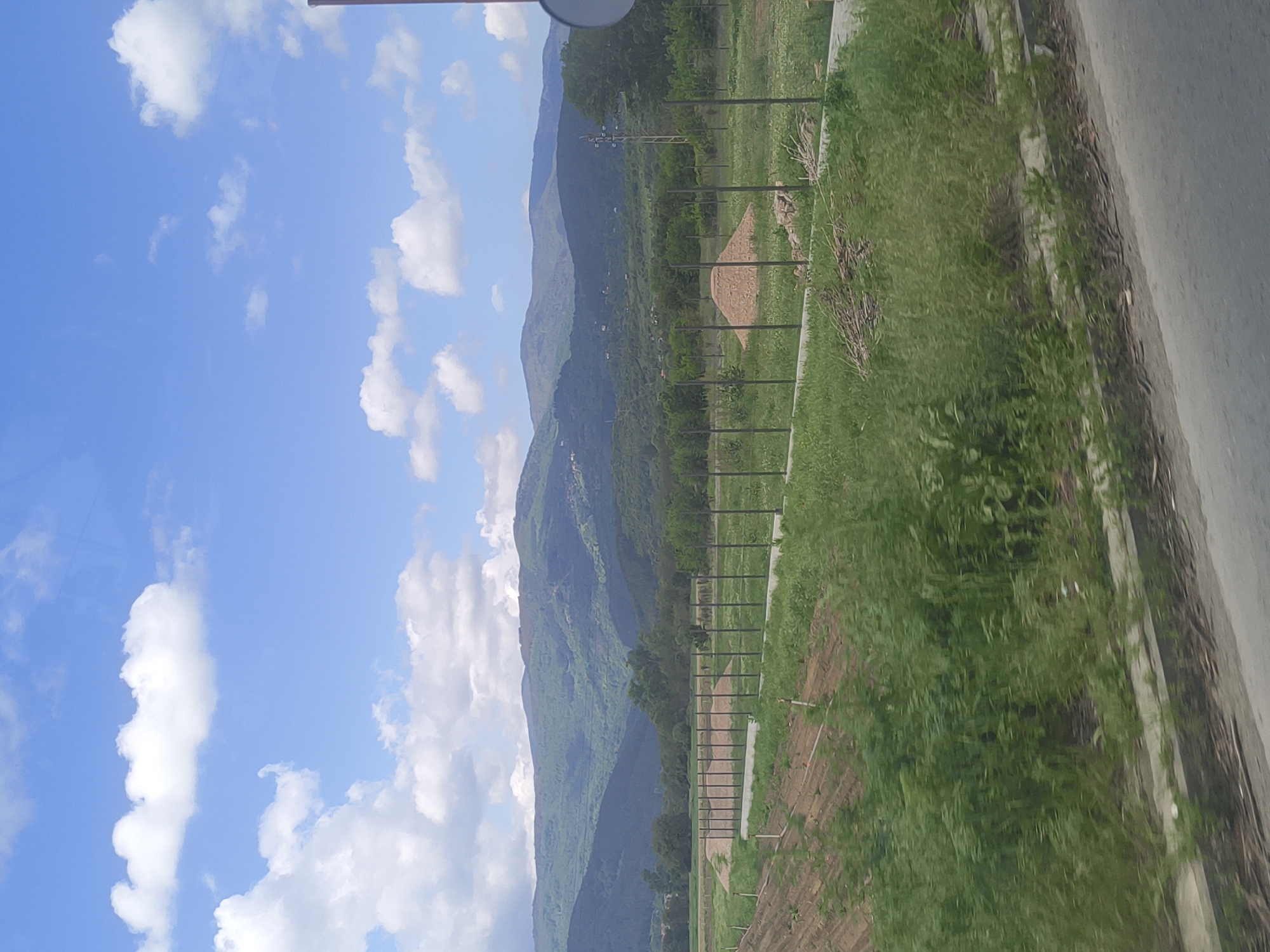The image shows a landscape in Piperkov Chiflik, Bulgaria. Mountains covered in green vegetation are visible in the background. A blue sky with white clouds is above the mountains. A fence runs along the right side of the image, separating a green field from a road. The road is made of grey concrete and runs along the right edge of the image. The photograph was taken facing the mountains, with the sky to the left.