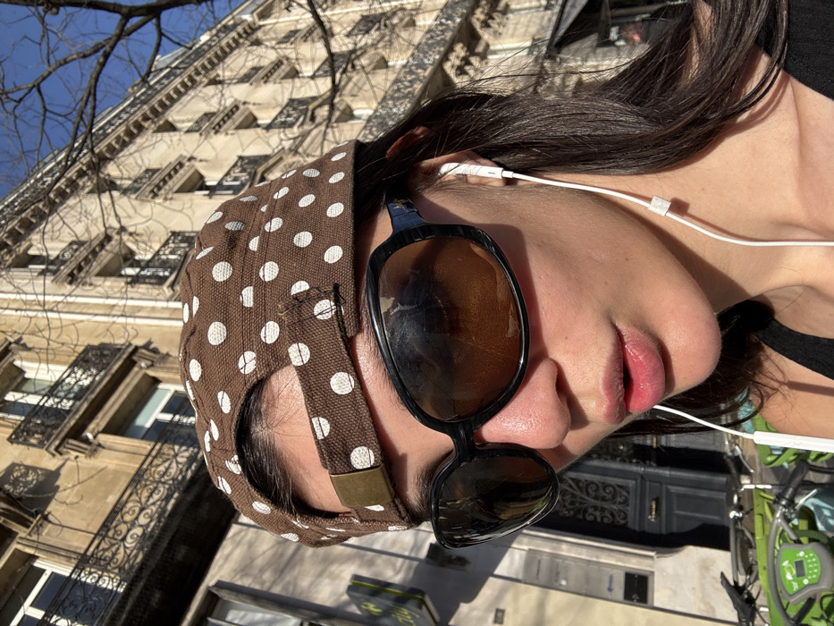 A close-up, outdoor shot depicts an individual in Paris, France, situated in front of a classic Haussmannian building. The person is partially obscured by the frame, with their face, neck, and shoulders visible. They wear a brown baseball cap with white polka dots, adjusted with a brass buckle, worn backward. Large, dark, oval-framed sunglasses cover their eyes, reflecting the sunny environment. White wired earbuds are inserted into both ears, with the cord extending downwards. The person's lips are slightly parted, and dark hair is visible beneath the cap and around the ears. In the background, to the left, a partial view of a dark, ornate building entrance is present, along with a bright green Vélib' Métropole bicycle docking station and several bicycles. To the right and above, a multi-story, light-colored Parisian building with intricate wrought-iron balconies and windows extends upwards. Bare tree branches are silhouetted against a clear blue sky, indicating bright, direct sunlight.