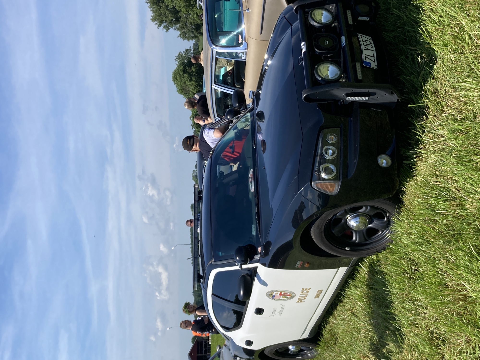 In Edenberga, Sweden, on a clear, sunny day, a black and white police car is parked on a grassy field. The car, a Dodge Charger, is the primary subject. It features a bull bar on the front with multiple lights, a visible license plate reading "ZLY 557", and the word "POLICE" emblazoned on its side in gold lettering. A circular police emblem is also present on the door. Several individuals are standing around the car and in the background, observing it or other vehicles. A tan vintage car is parked to the right of the police car. The environment is a grassy outdoor area with trees and buildings visible in the distance under a blue sky with scattered clouds.