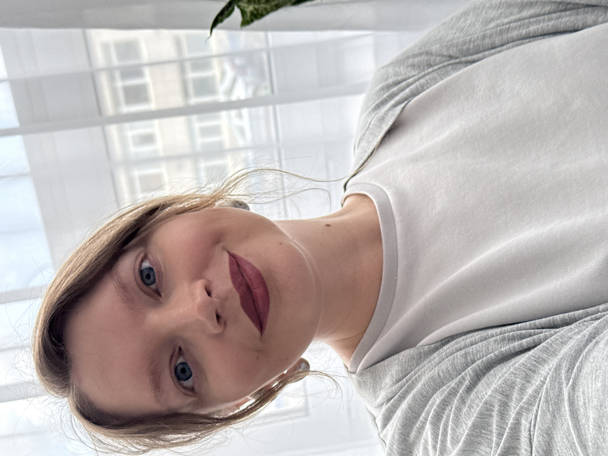 A woman with fair skin, blue eyes, and auburn hair is looking down at the camera. She is wearing a white t-shirt with gray sleeves. Her lips are painted with a dark red lipstick. The background is blurred, but appears to be a window with white blinds. There is a potted plant visible in the upper right corner. The photo was not taken in Izhevsk, Russia.