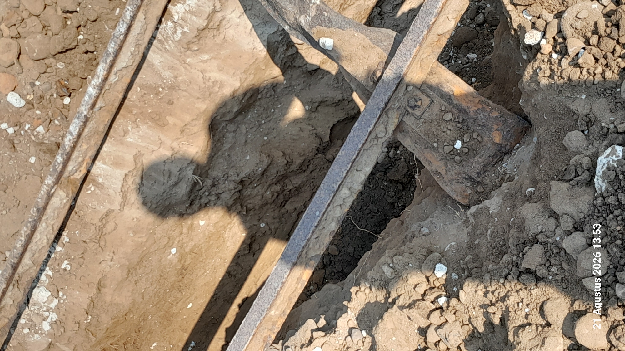 A shadow of a person is cast onto the dirt walls of a trench. The trench appears to be under construction or excavation, with exposed soil and rocks. Several pieces of metal, likely construction materials or tools, are visible within the trench. The overall scene suggests an outdoor, possibly rural or construction site environment. The date and time stamp "21 Agustus 2026 13.53" is visible in the bottom left corner of the image. The location is identified as Takeran, Indonesia.