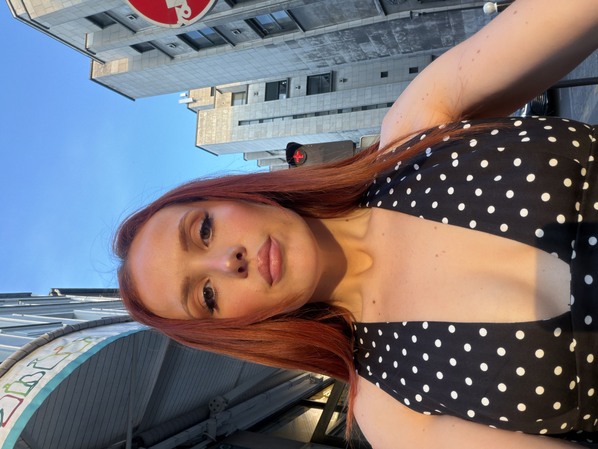 A selfie of a fair-skinned woman with reddish-brown hair wearing a black and white polka dot dress. The photograph appears to be taken outdoors in a city, evidenced by buildings and street infrastructure in the background. The blue sky is visible in the upper portion of the image. The buildings have a modernist architectural style. The location is Paris, France, indicated by the red and white "sens interdit" (no entry) sign visible atop one of the buildings.