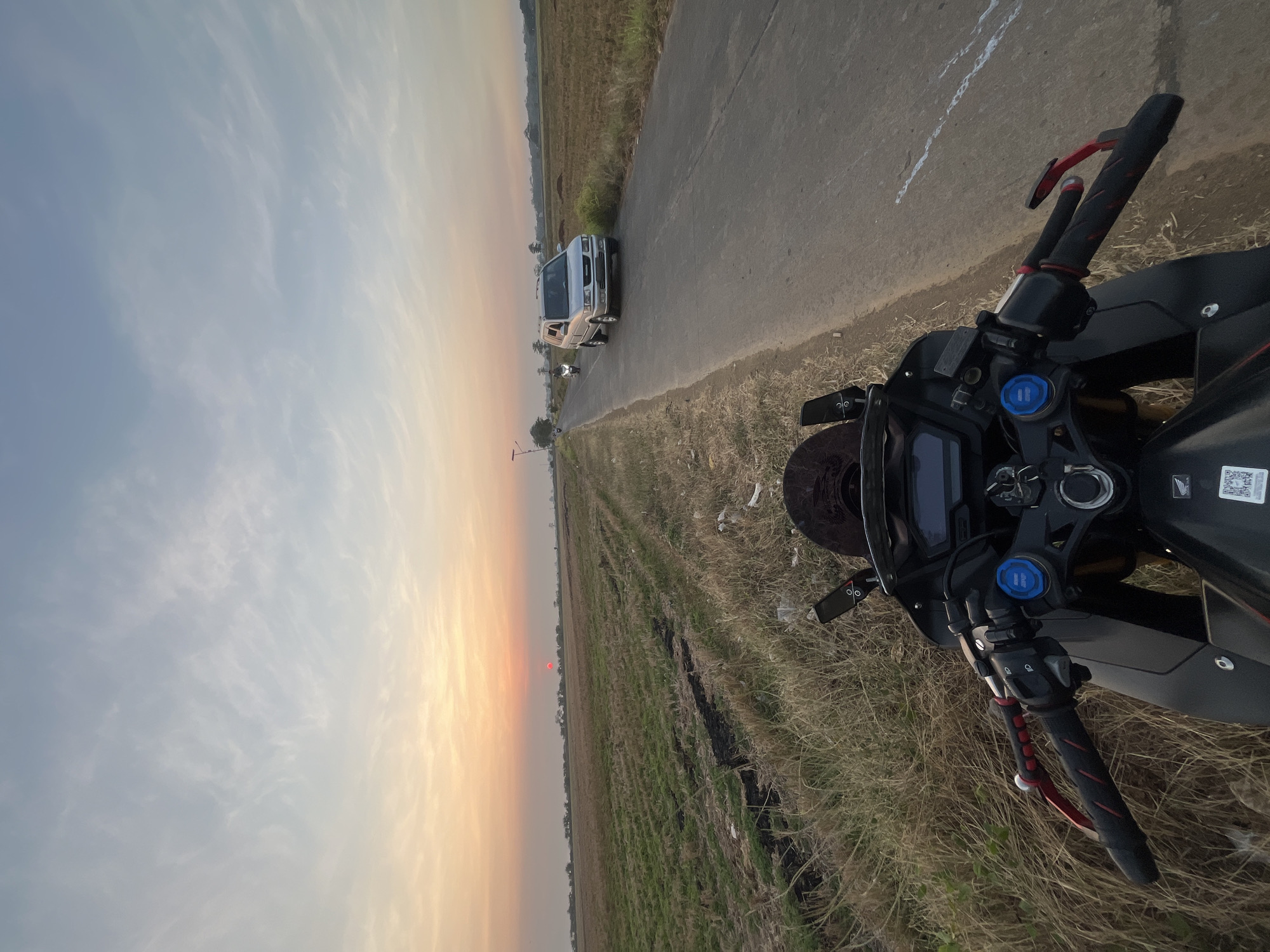 The image captures a motorcycle's perspective on a rural road at sunset in Pecangaan, Indonesia. The foreground features the handlebars and dashboard of a black motorcycle. Beyond the motorcycle, a paved road stretches into the distance, with a silver SUV and a motorcycle driving on it. The road is bordered by dry, grassy fields. In the background, a hazy orange and pink sunset illuminates the sky with wispy clouds. The overall scene depicts a serene, rural landscape during the golden hour.