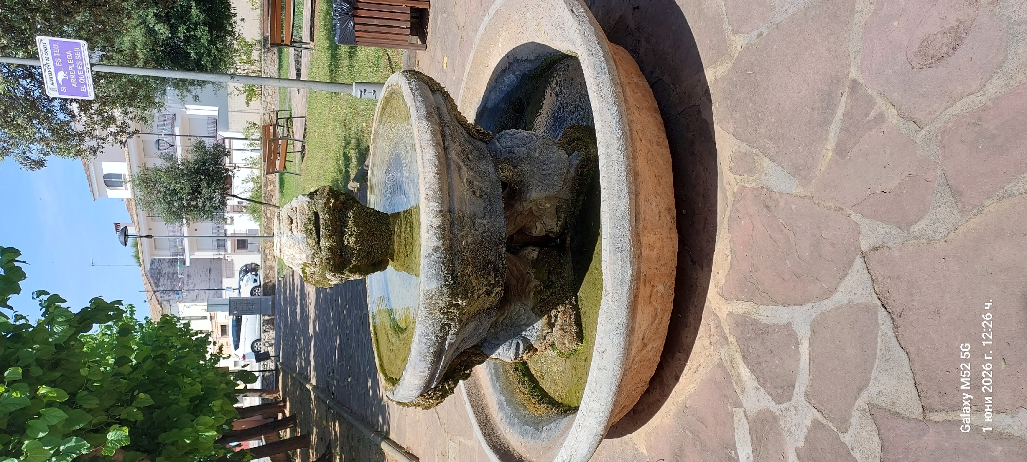The image shows a stone fountain in a park in the city of Canals, Spain. The fountain is made of weathered stone and has multiple tiers with water collecting in the basins. The surrounding area is paved with flagstones. A sign indicating the "AJUNTAMENT DE CANALS" is visible in the background, and the text on the sign reads "SI ES TEU, ARREPLEGA EL QUE ES SEU." The image was taken on June 1, 2026, at 12:26 p.m., as indicated by the date and timestamp in the lower right corner. The camera used was a Galaxy M52 5G.
