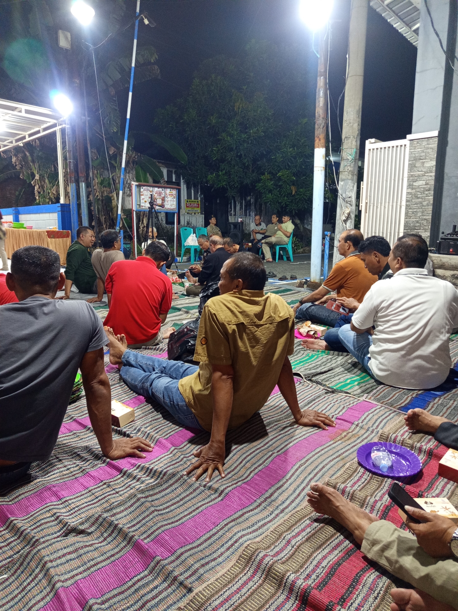 A group of men are gathered outdoors at night in Masangan Kulon, Indonesia, seated on colorful striped mats. They appear to be attending an event or gathering, possibly a ceremony or social function. Several men are looking towards a tripod-mounted camera or projector in the background, suggesting a presentation or recording is taking place. The lighting is provided by bright overhead lamps, illuminating the scene and creating shadows. Some individuals are engaged in conversation, while others are observing. Objects visible include small food packages, a purple plate with a bottle of water, and a mobile phone. The environment features trees and buildings, indicating a residential or community area. The overall atmosphere seems relaxed and communal.