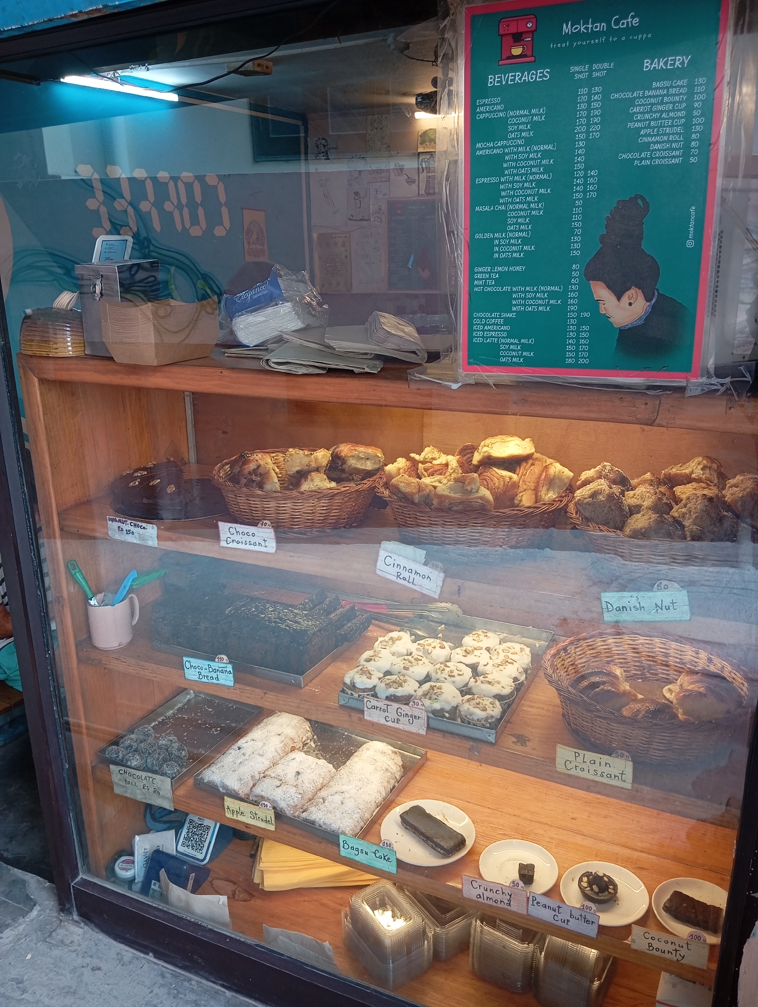 The image shows a bakery display case in Jonk, India. The case has three shelves filled with various baked goods.
Top shelf: Walnut chocolate cake, chocolate croissants, cinnamon rolls, and Danish nuts.
Middle shelf: Chocolate banana bread, an assortment of items labeled as chocolate balls, apple strudel, and carrot ginger cups.
Bottom shelf: Bagsu cake, items labeled as crunchy almond, peanut butter cups, and coconut bounty.
A menu is attached to the side of the display case, listing beverages and bakery items with prices. A cash register and other items are visible on the counter above the display case.