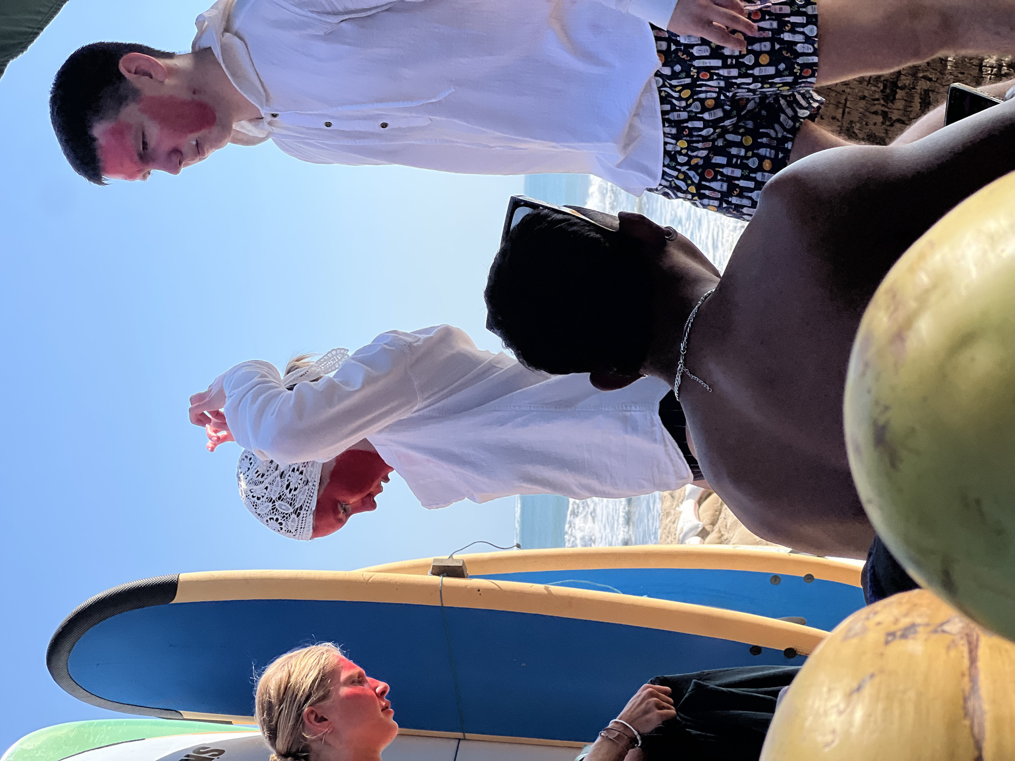 The image depicts a sunny outdoor scene on a beach, likely in Kuta, Indonesia, as evidenced by the presence of surfboards and the ocean. In the foreground, two coconuts are partially visible. Several individuals are present, all appearing to have red face paint on.
On the left, a person with blonde hair tied back is seen from the side, facing down. Next to them are two surfboards, one blue and yellow, and another with white and green elements.
In the center, a person wearing a white headscarf and a white long-sleeved shirt is adjusting their headscarf. Their face is covered in red paint.
To the right, a young person in a white hooded shirt and patterned shorts with cocktail imagery is looking off to the side. Their face also has red paint.
In the mid-ground, the back of a person is visible, wearing a silver chain necklace and dark shorts. This person is looking towards the ocean. The ocean with waves is in the background, and the sky is clear and blue. The sand and some white fabric are visible on the beach.