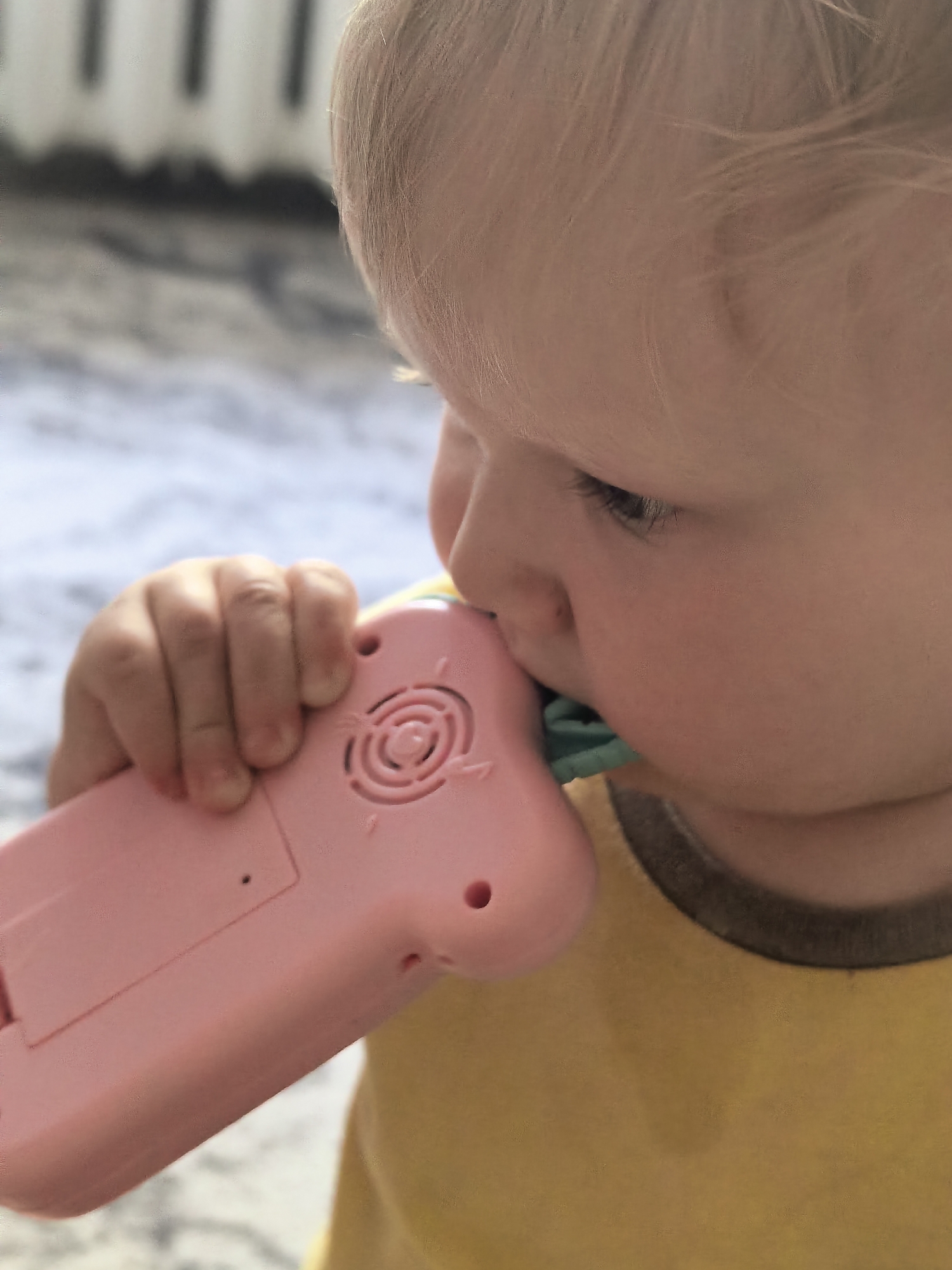 A close-up, slightly angled shot shows a toddler holding a pink toy phone to their mouth. The toddler's face is partially visible, with their eyes looking down and their blonde hair catching the light. Their small hand is wrapped around the toy phone, which has a circular speaker grill and what appears to be a green button or attachment near the toddler's mouth. The background is blurred, suggesting an outdoor or lightly lit indoor setting, with vertical elements in the far distance that could be a fence or railing. The toddler is wearing a yellow shirt with a brown collar. The image does not contain any elements that would indicate a specific location within the city of Sterlitamak, Russia.