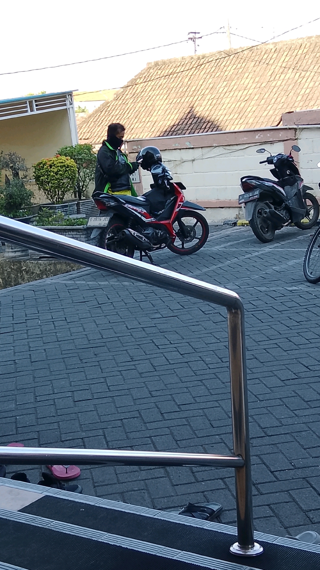 A person wearing a black and green jacket and a black face mask is standing next to a red motorcycle in what appears to be a parking area in Ngipik, Indonesia. The person is holding a black helmet, seemingly placing it on the motorcycle. Another black motorcycle is parked to the right of the red one, and a portion of a bicycle is visible on the far right. The ground is paved with dark gray bricks, and a metal railing runs across the foreground. In the background, a building with a tiled roof and a portion of a light-colored wall are visible. Several pairs of flip-flops are scattered on the paved ground near some stairs in the lower part of the image.