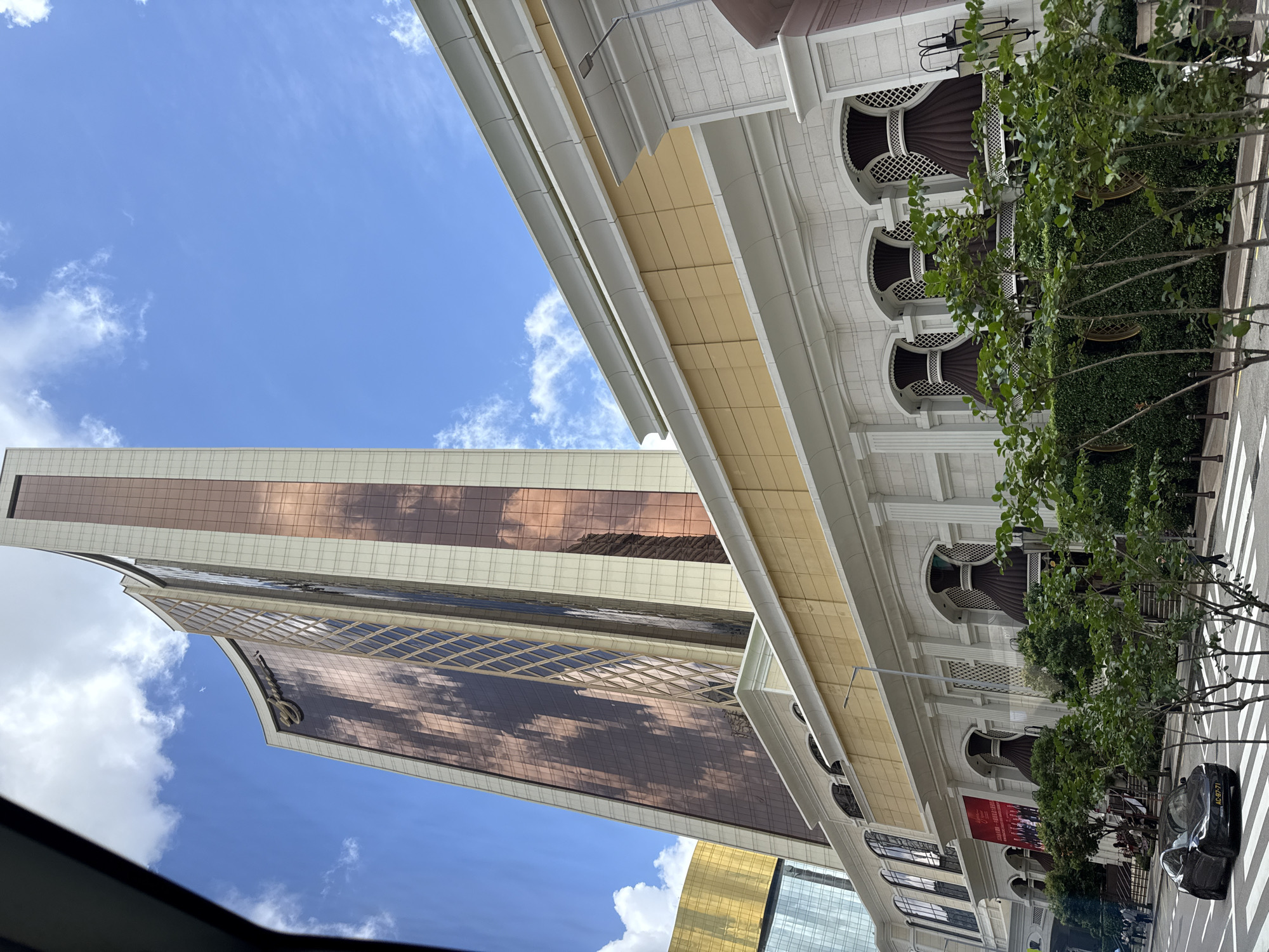 This is a low-angle, outdoor, daytime, eye-level shot of the Grand Lisboa Palace Resort Macau. The resort's iconic, curved, rose-gold and cream-colored towers dominate the upper portion of the frame, reflecting the blue sky and white clouds. Below the towers, a section of the resort's facade is visible, featuring a light yellow band above a white, classical-style base. This lower section has arched windows with decorative, dark brown enclosures and ornate white detailing. Green bushes and trees are in the foreground, partially obscuring the lower facade. A black car is in the left lane of the street, with a pedestrian crossing in front of it. The street is paved, and there are sidewalks and streetlights visible. The overall scene depicts a modern luxury resort in an urban setting on a clear day.