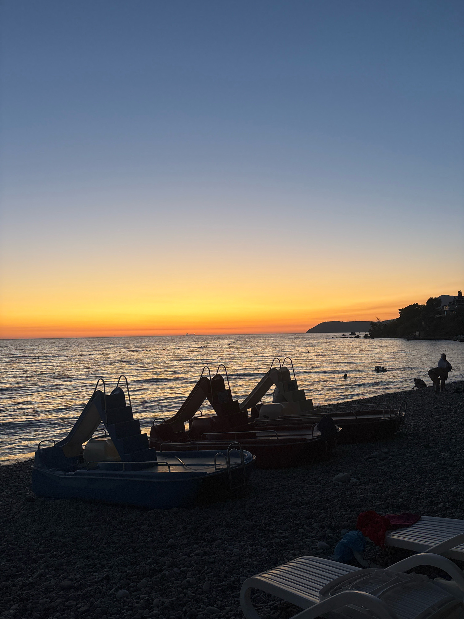 This image depicts a serene beach scene at sunset in Bar, Montenegro. The foreground features several brightly colored pedal boats lined up on a pebble beach. Behind the boats, the calm sea stretches to the horizon, reflecting the warm hues of the setting sun. In the distance, a silhouette of hills marks the coastline. A few individuals are visible on the beach and in the water, enjoying the tranquil evening. The sky transitions from a deep blue at the top to vibrant oranges and yellows near the horizon, indicating the end of the day.