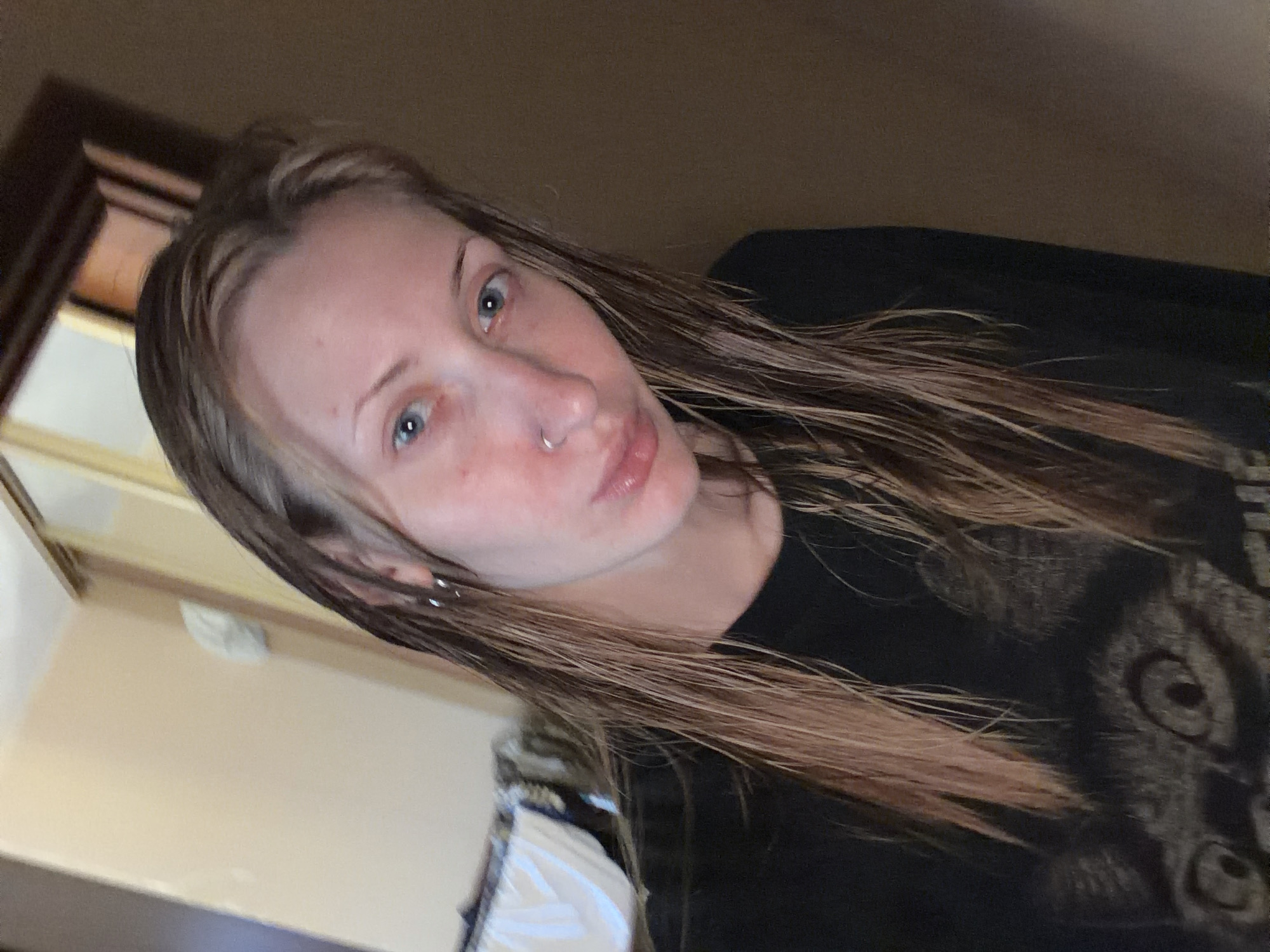 The media file is a close-up, angled selfie of a person. The subject appears to be a young woman with light skin, light blue eyes, and medium-length, light brown hair that is slightly wet. She is wearing a black t-shirt with a graphic design of an animal's face, possibly a wolf or a cat, on the front. She has a silver hoop nose ring and small silver hoop earrings in her visible ear. Her facial expression is neutral with slightly pursed lips. The background is blurry and appears to be an indoor setting with neutral-toned walls and some wooden furniture. The lighting suggests it could be daytime, but the indoor setting makes it difficult to determine. There is no visible text in the image. The overall mood is casual and personal.