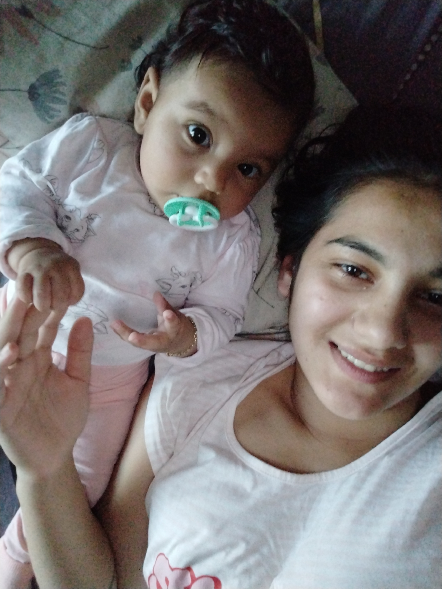 A selfie shows an adult woman and a baby lying on bedding. The baby, wearing a light pink outfit and holding a pacifier, is positioned near the woman's chest. The woman, wearing a white t-shirt, smiles at the camera and holds out her hand. The background includes a pillow with a flower pattern. The location is unknown.