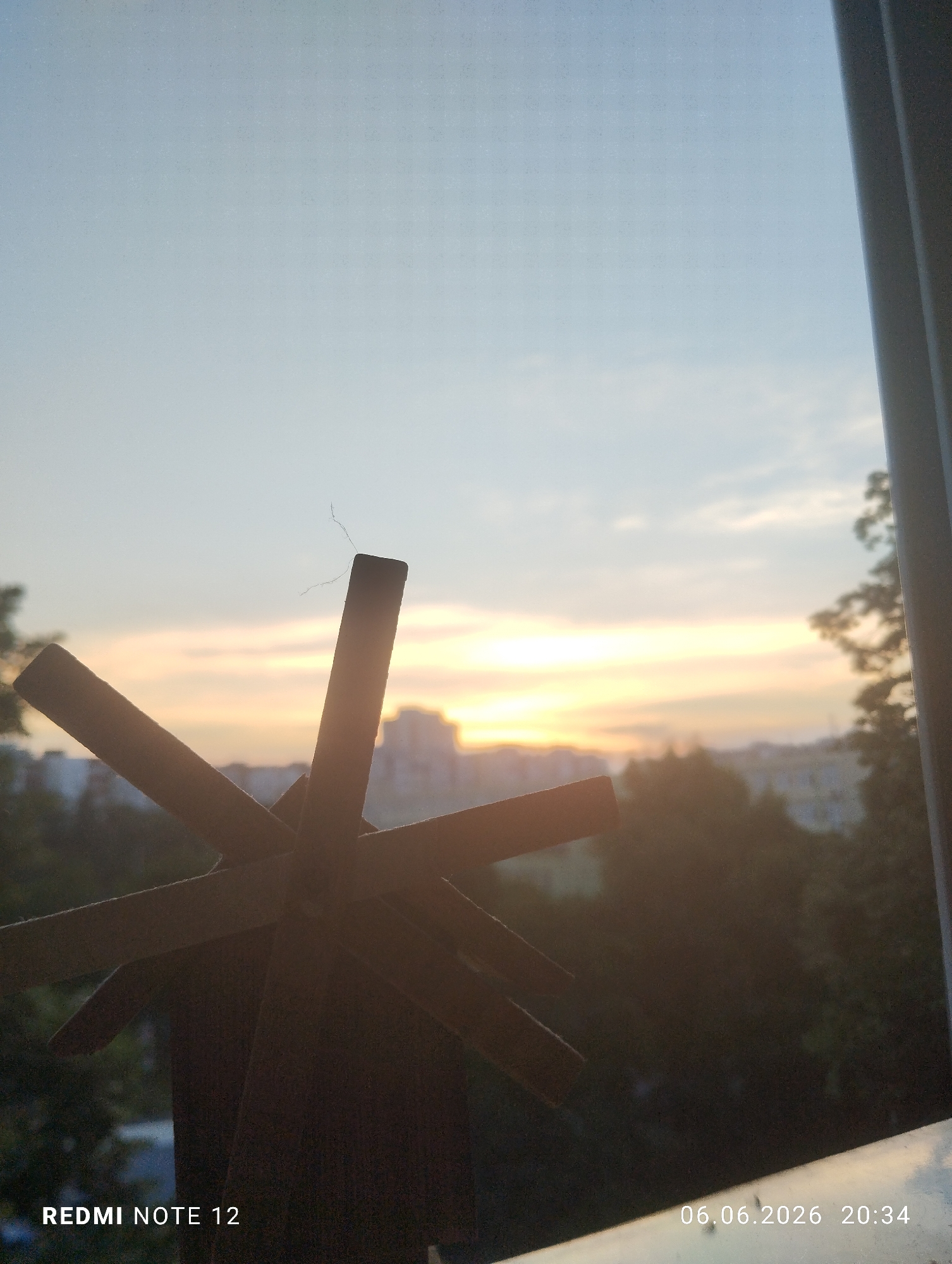 The image displays an urban sunset scene on June 6, 2026, at 20:34, in what is contextually specified as Burgas, Bulgaria. In the immediate foreground, a dark, silhouetted object, resembling a decorative wooden windmill or a stylized pinwheel, occupies the lower left and central area. Four distinct blades are visible, with a small, wispy, light-colored fiber or hair adhering to the tip of the uppermost blade. To the right, a dark, vertical structural element, consistent with a window frame or balcony railing, frames the scene. The mid-ground reveals a dark, silhouetted urban landscape, featuring multiple building structures and a dense canopy of trees. The background consists of a gradient sky, transitioning from a pale blue at the top to warm orange and yellow tones at the horizon, indicative of a sunset. No human figures or discernible actions involving people are present. The image bears embedded text "REDMI NOTE 12" in the bottom left and the timestamp "06.06.2026 20:34" in the bottom right.