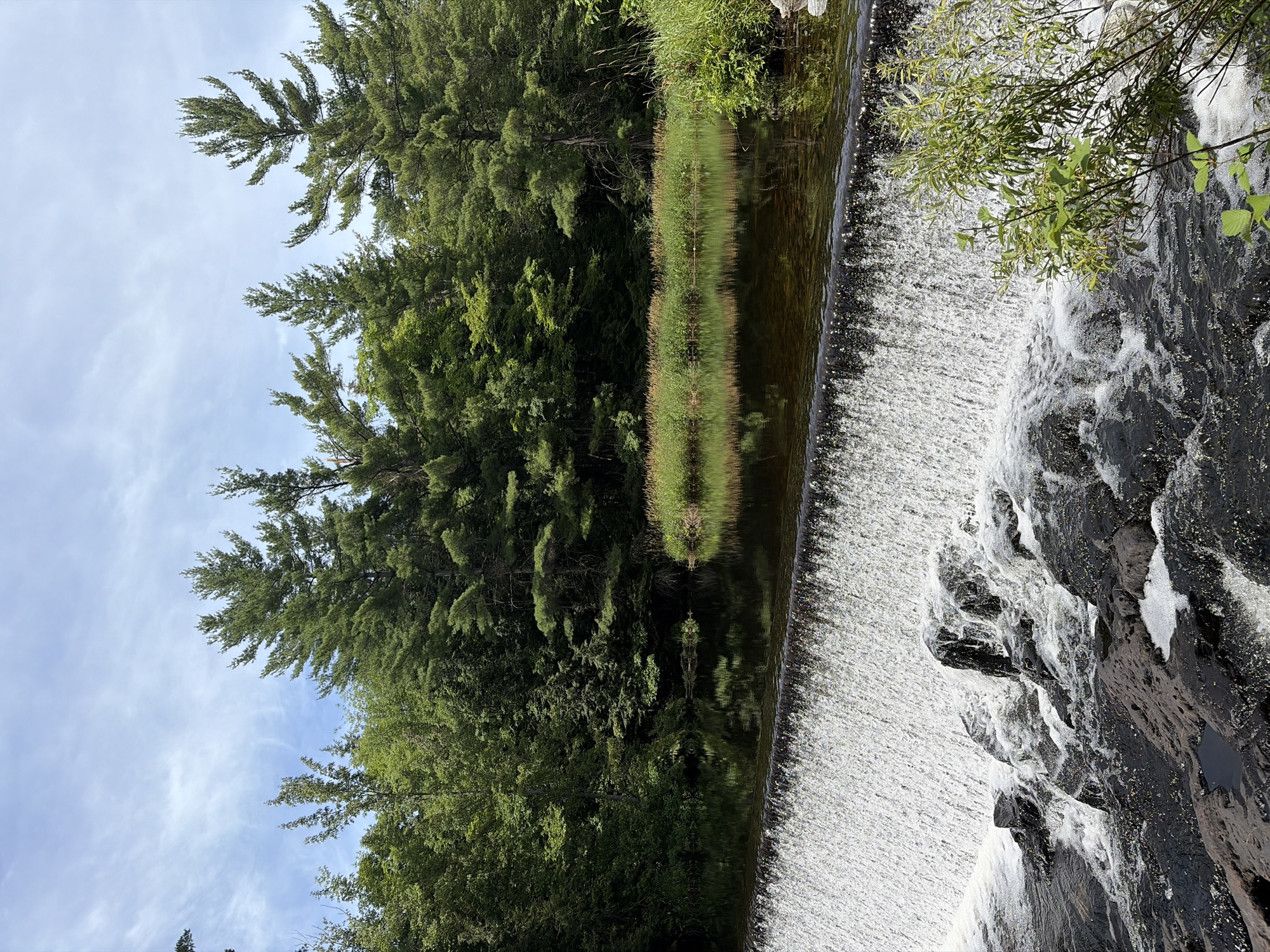 This image captures a natural scene in Addington Highlands, Canada, featuring a wide, low waterfall cascading over dark rocks. The water is frothy at the base of the falls. Above the waterfall, a calm body of dark water reflects the surrounding environment. The background is dominated by a dense forest of evergreen trees and other lush green foliage. The sky above is partly cloudy with wisps of white clouds against a light blue expanse. The overall impression is one of a serene, natural landscape.