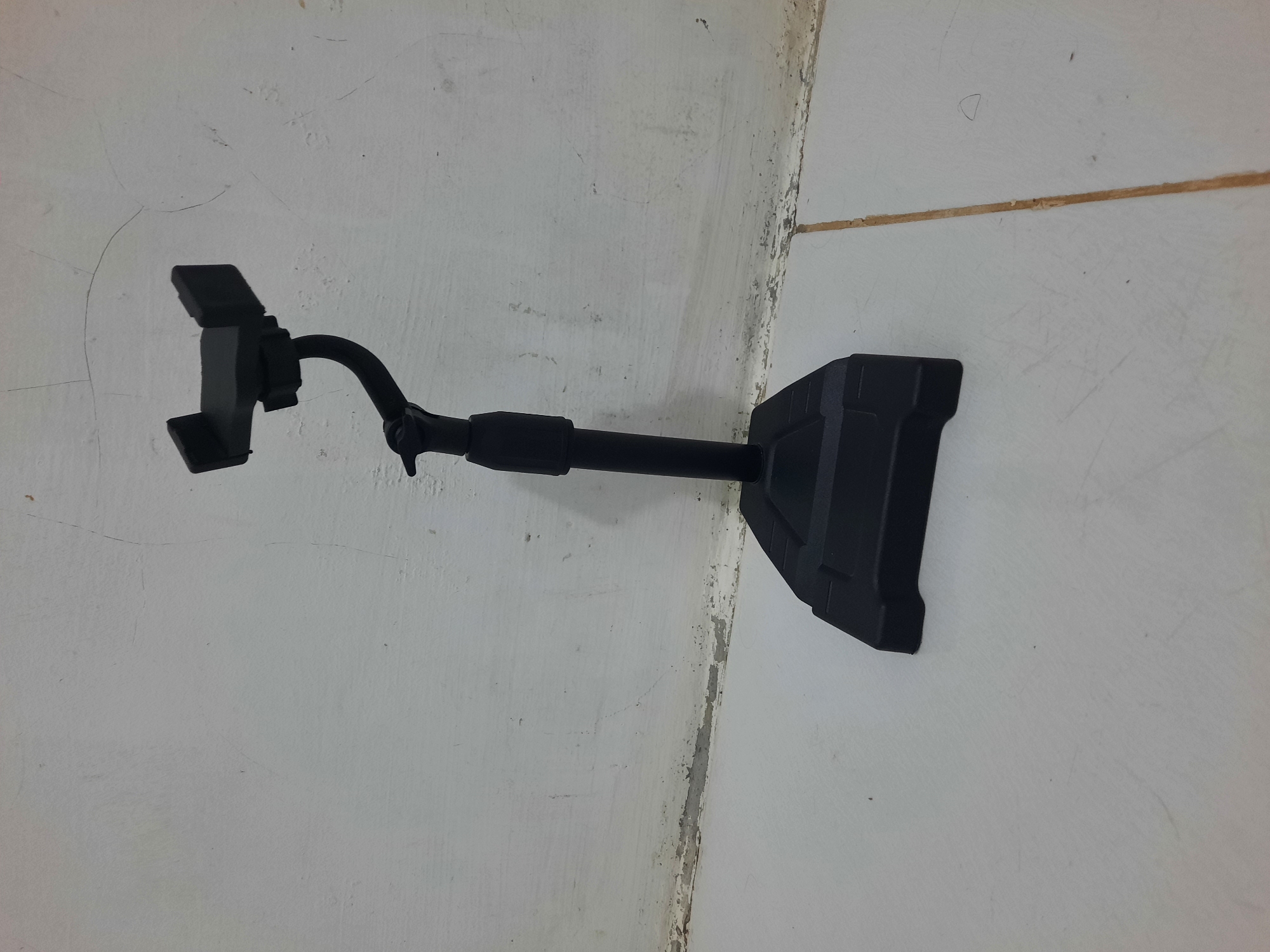 A black cell phone stand is pictured against a white, textured wall and a light-colored tiled floor. The stand has a wide, flat base and a vertical pole that supports a flexible gooseneck arm. The arm ends in a clamp designed to hold a smartphone. The wall shows signs of age and wear, with visible cracks and peeling paint near the floor. The tiled floor has visible grout lines. The scene is well-lit, with no discernible shadows. The setting is an interior space, likely a room or an outdoor covered area with a plain wall and floor. The image does not contain any people or discernible elements that would link it to Lhokseumawe, Indonesia.