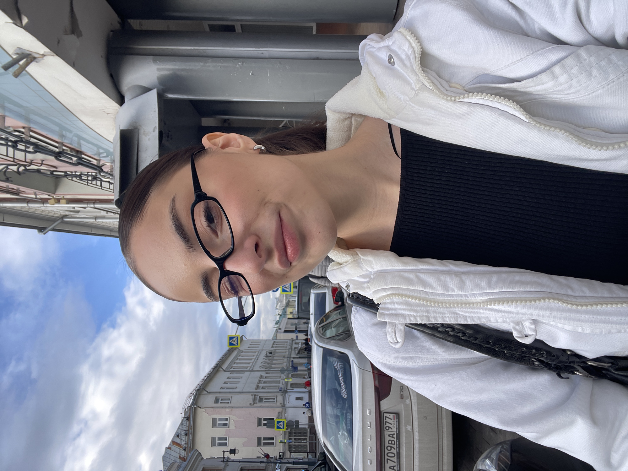 A medium-close-up shot depicts a young woman with fair skin, wearing black-framed glasses. She is dressed in a black ribbed top and a white jacket with a zipper. A black shoulder bag is visible. The background shows a city street scene, with buildings, pedestrian crosswalks, street signs, a cloudy sky, and a white car with license plate A709BA 977. The car is labeled as Exeed. The scene is set in Moscow, Russia, identifiable through the license plate.