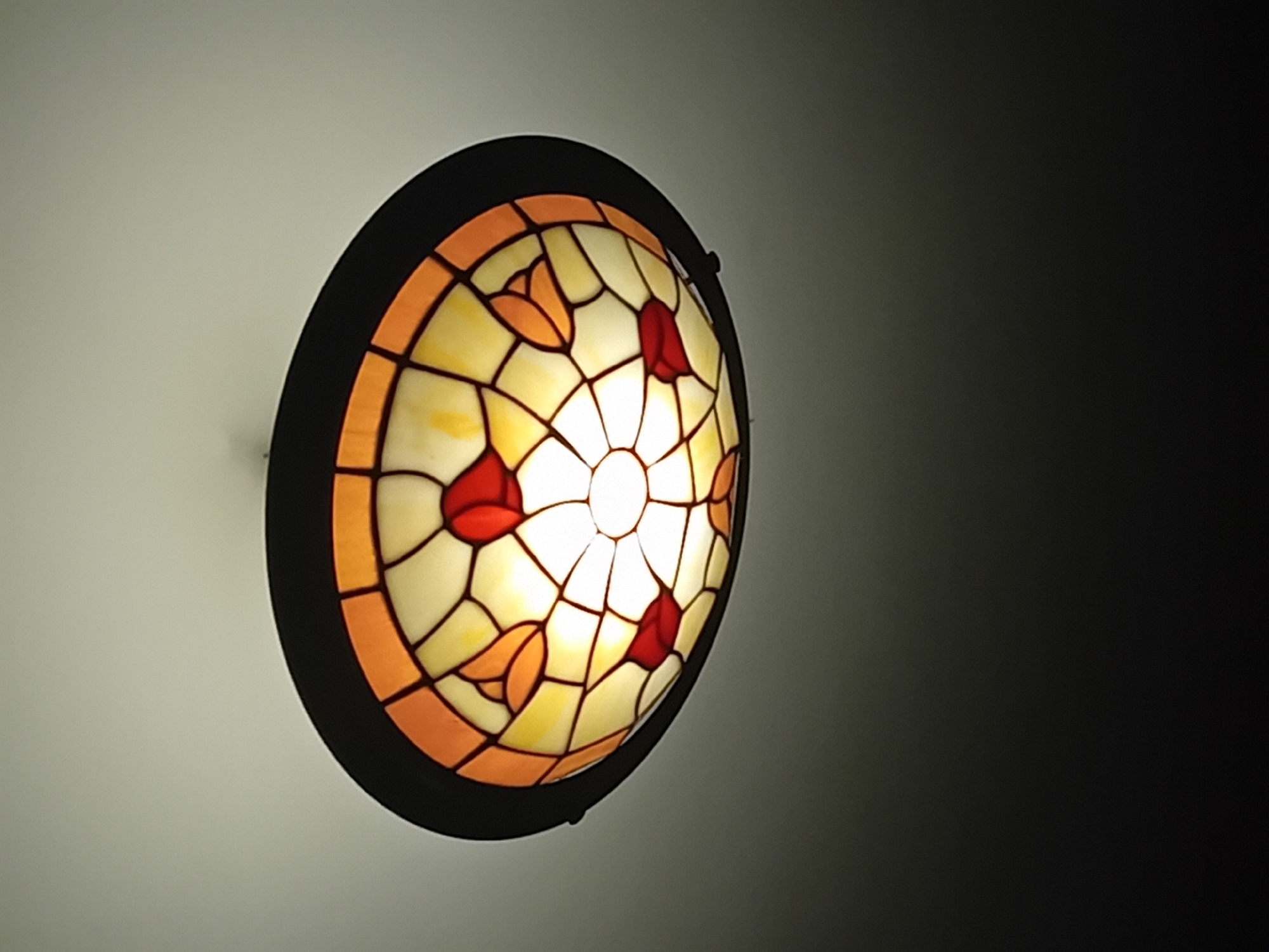 A brightly illuminated stained-glass lamp with a black metal frame is mounted on a wall. The lamp's design features off-white glass with orange and red accents. The light source within the lamp is bright and centered. The wall behind the lamp is two-toned, transitioning from a lighter shade to a darker shade as it moves away from the lamp. The city and location are unknown.