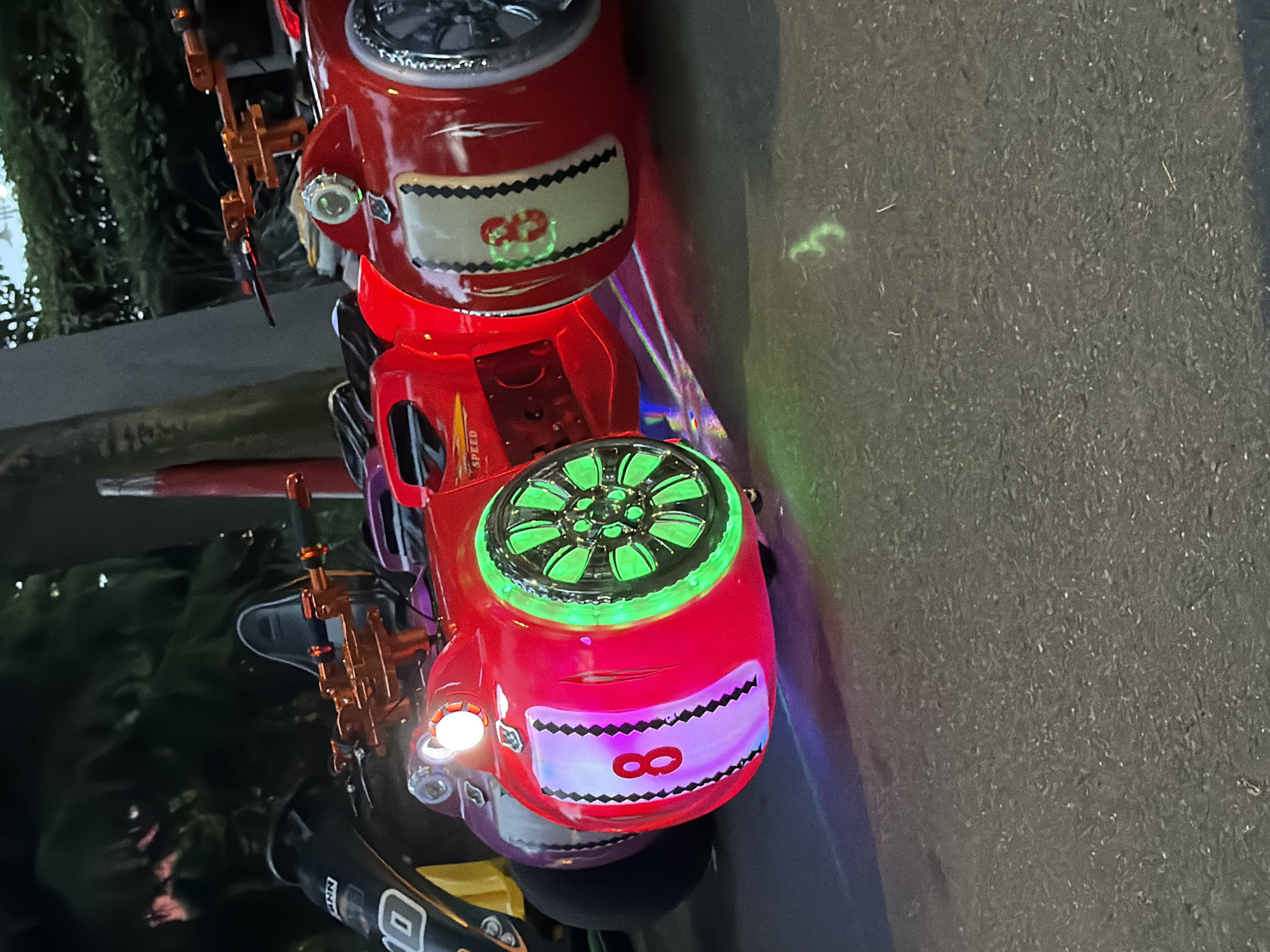 The image shows a close-up, vertically oriented view of two red, toy electric vehicles, likely for children. The vehicles are detailed with chrome accents, headlights, and colorful lights. The front vehicle has a large, green, illuminated wheel with a fan-like design. It also features a decorative panel with a black and white zig-zag border and a large red number "8" within a purple illuminated area. The vehicle behind it appears similar, with a white panel displaying a red number "88". The vehicles are positioned on an asphalt surface, possibly a street or walkway. In the background, dark green foliage is visible, suggesting an outdoor setting. The lighting indicates it is either dusk or night, with artificial lights from the vehicles creating colorful reflections on the ground. The overall impression is of a fun, possibly commercial, amusement ride. No people are visible in the frame. The location context provided is Burgas, Bulgaria, but there are no specific visual cues in the image to confirm this.