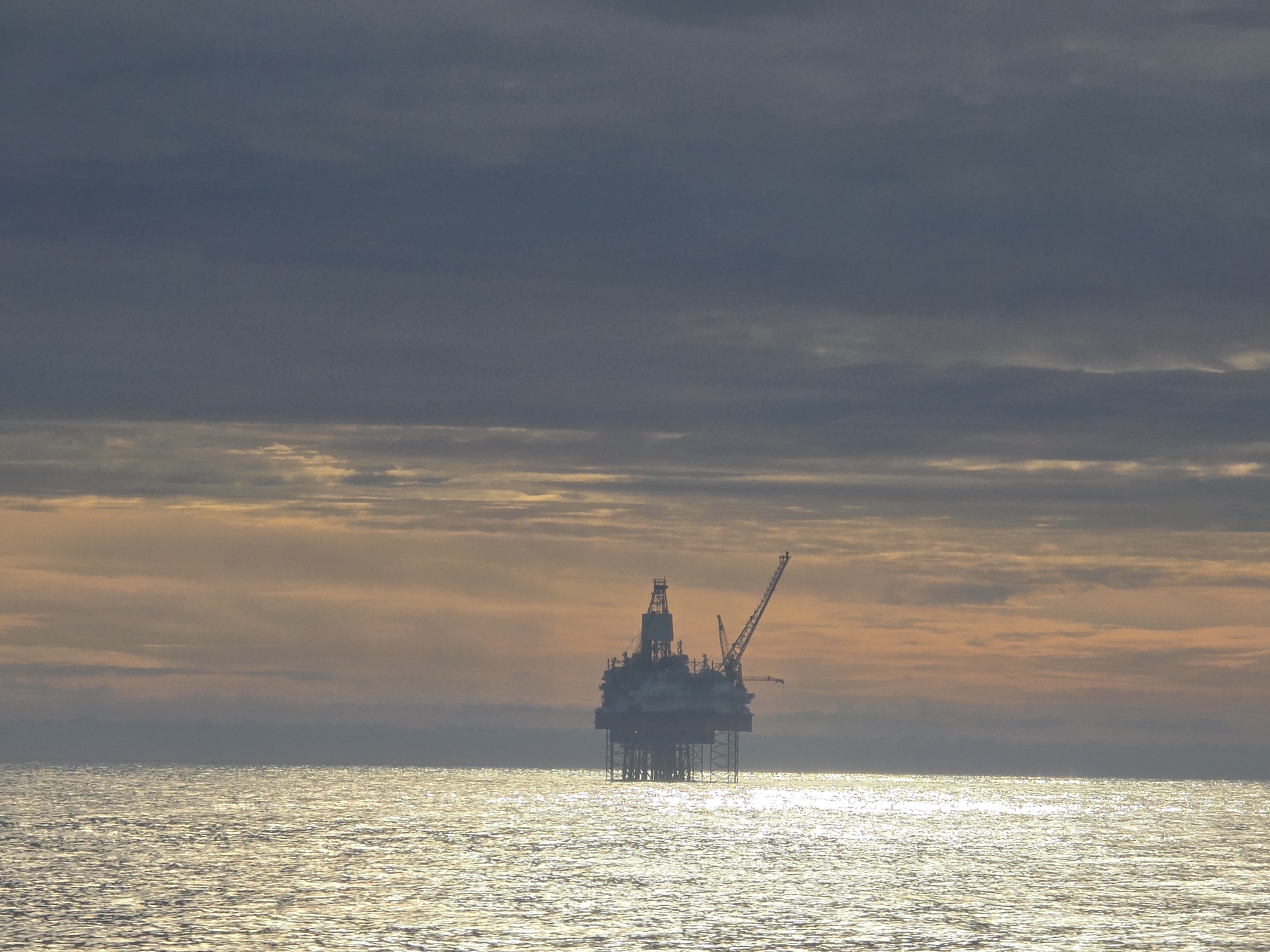 An oil rig stands on a flat, reflective body of water, likely an ocean, under a sky with layers of clouds. The upper part of the sky consists of dark, gray clouds, while the lower portion has a mix of lighter clouds with hints of golden color, suggesting either sunrise or sunset. The oil rig is a silhouette against the lighter sky, and its structure is somewhat visible, including a crane. The water reflects the golden light, creating a shimmering effect.