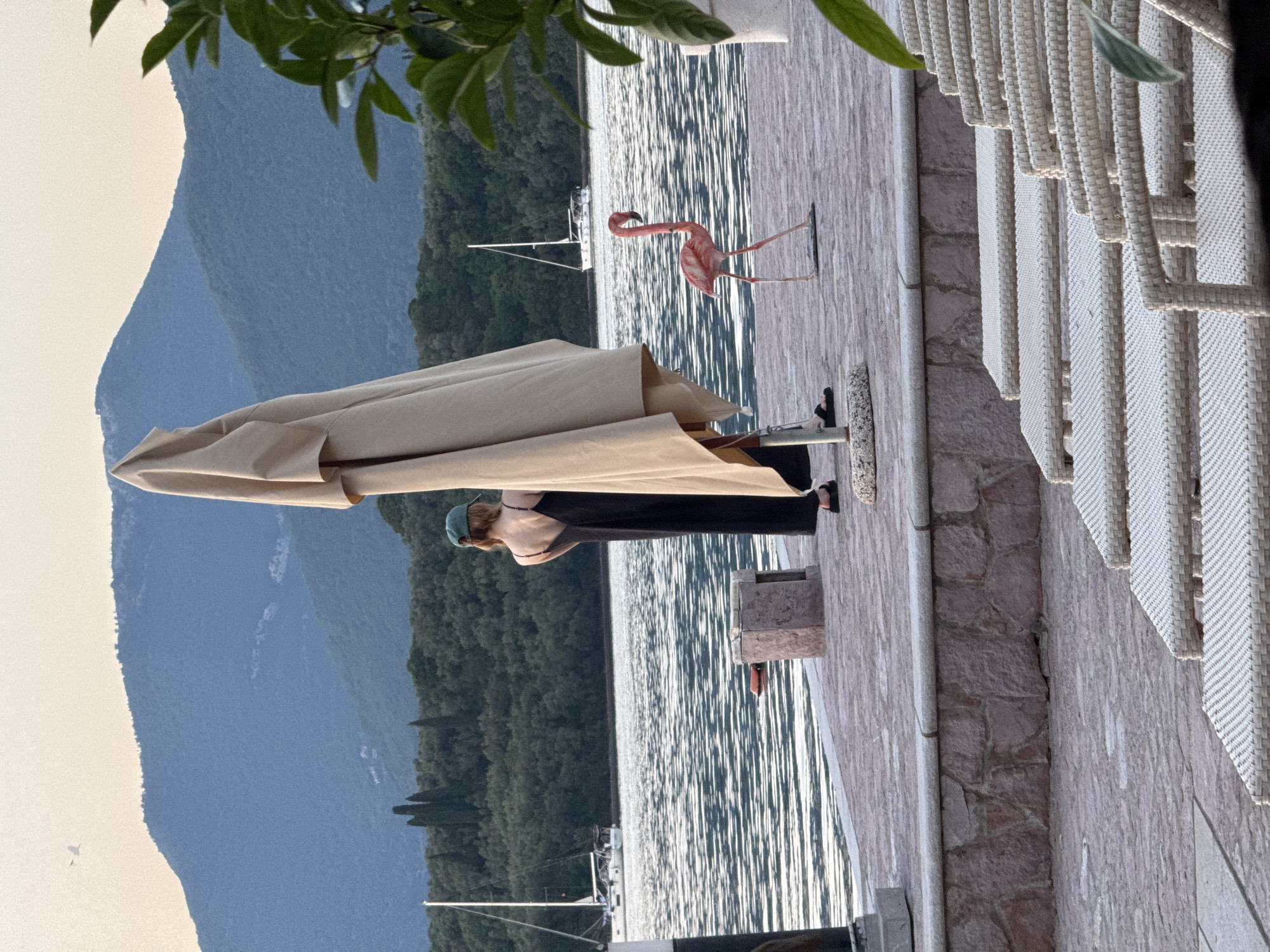 The image depicts a scene from Bogišići, Montenegro. A woman in a black dress and a green cap stands under a tan umbrella, overlooking the water. To her right, there is a pink flamingo statue on a wooden platform next to wicker chairs. Behind her, mountains and a forest line the horizon. A sailboat is visible on the water. The setting appears to be a patio or terrace along the waterfront.