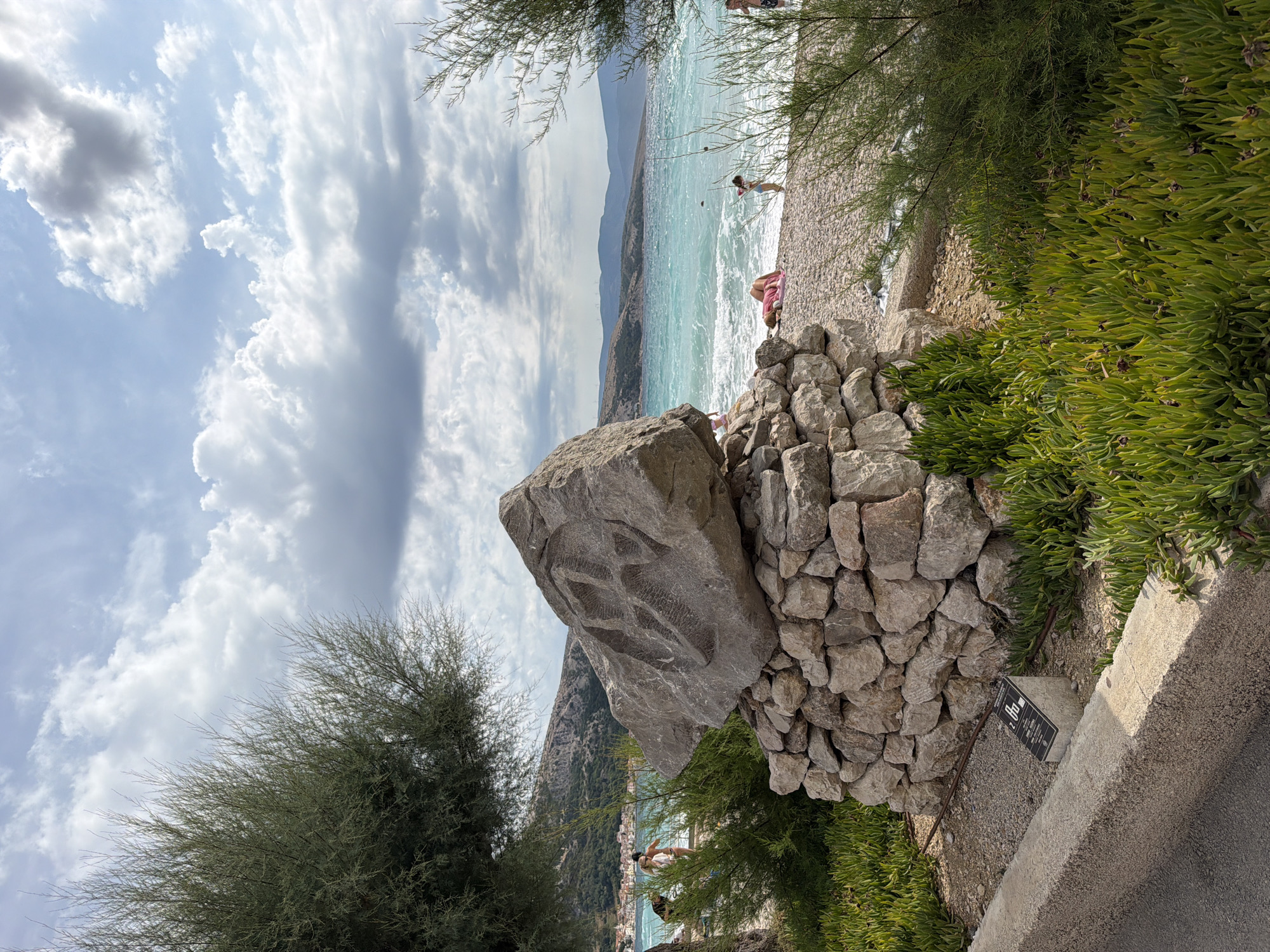 This is a full outdoor, eye-level shot taken on a slightly cloudy day at the beach in Općina Baška, Croatia. In the foreground, a large, rough stone sculpture sits atop a foundation of stacked stones. The sculpture is carved with an abstract pattern. Green, succulent-like plants grow around the base of the sculpture and a concrete curb runs along the left side of the frame. Behind the sculpture, a pebbled beach stretches towards the turquoise sea. Several people are visible on the beach, some wading in the water, others sunbathing or walking. Trees with wispy branches are on either side of the frame, framing the view of the beach and the distant, hazy hills. A small, dark plaque with white text is visible at the base of the stone foundation, to the left of the sculpture.