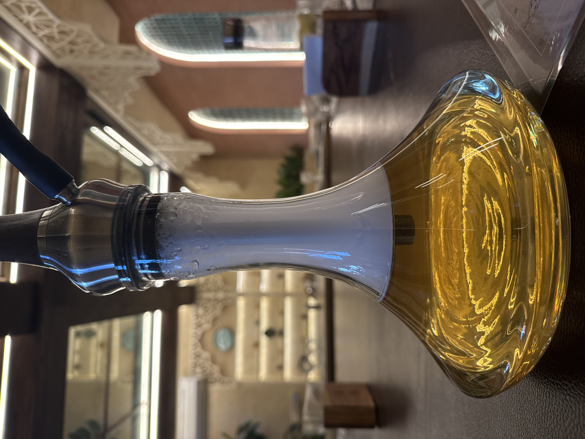 The image captures a hookah, centrally positioned and angled towards the upper right, against a background suggesting a restaurant or lounge interior. The hookah has a black hose connected to a metallic body, with a translucent midsection filled with a smoky substance. The base is a bulbous, amber-colored glass. The background features interior design elements such as rounded ceiling recesses with embedded lighting, along with decorative molding. The location is North Kuta, Indonesia.