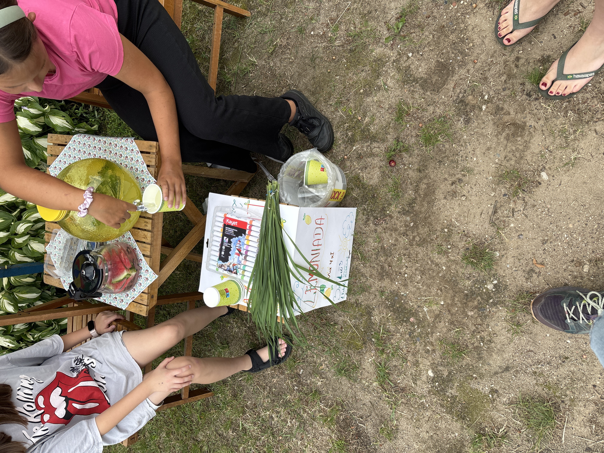 In Trześcianka, Poland, two young females are seated on wooden chairs at a wooden table outdoors. One female, wearing a grey t-shirt with a Rolling Stones logo, sits on the left. The other female, in a pink t-shirt and black pants, is on the right, pouring a clear yellow liquid from a large glass dispenser into a small green paper cup. On the table are a glass pitcher filled with watermelon slices, a floral-patterned cloth, and a box of markers. A box labeled "Lemoniada" with drawings is on the ground next to a bunch of long green grass. The ground is covered with dirt and sparse grass. In the lower portion of the image, the feet of two individuals wearing sneakers and flip-flops respectively are visible. The scene is set in a natural, grassy environment.